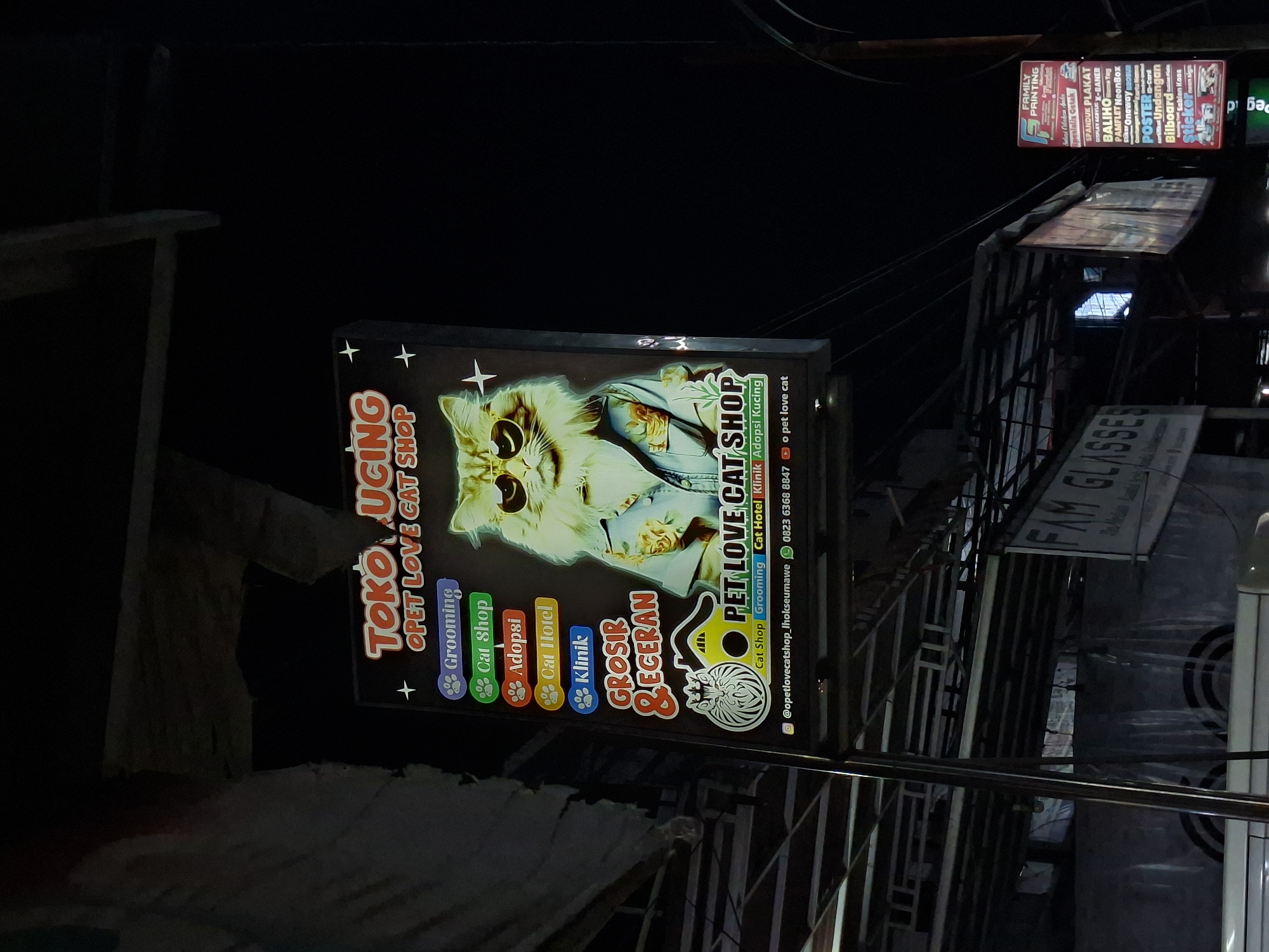 The image shows a night scene in Lhokseumawe, Indonesia. The focus is on a brightly lit sign for "Toko Kucing Opet Love Cat Shop," a pet store specializing in cats. The sign features a cartoonish image of a cat wearing sunglasses and a suit. Below the image, the sign lists services: grooming, cat shop, adoption, cat hotel, and clinic. The sign also displays contact information, including an Instagram handle and a phone number. In the background, another sign for "Family Printing" is visible, along with other commercial signs, indicating a business district. The scene is dark, with artificial lighting illuminating the signs and surrounding structures. There is no human presence in the image. The signs are mounted on a pole and wiring that are visible overhead.