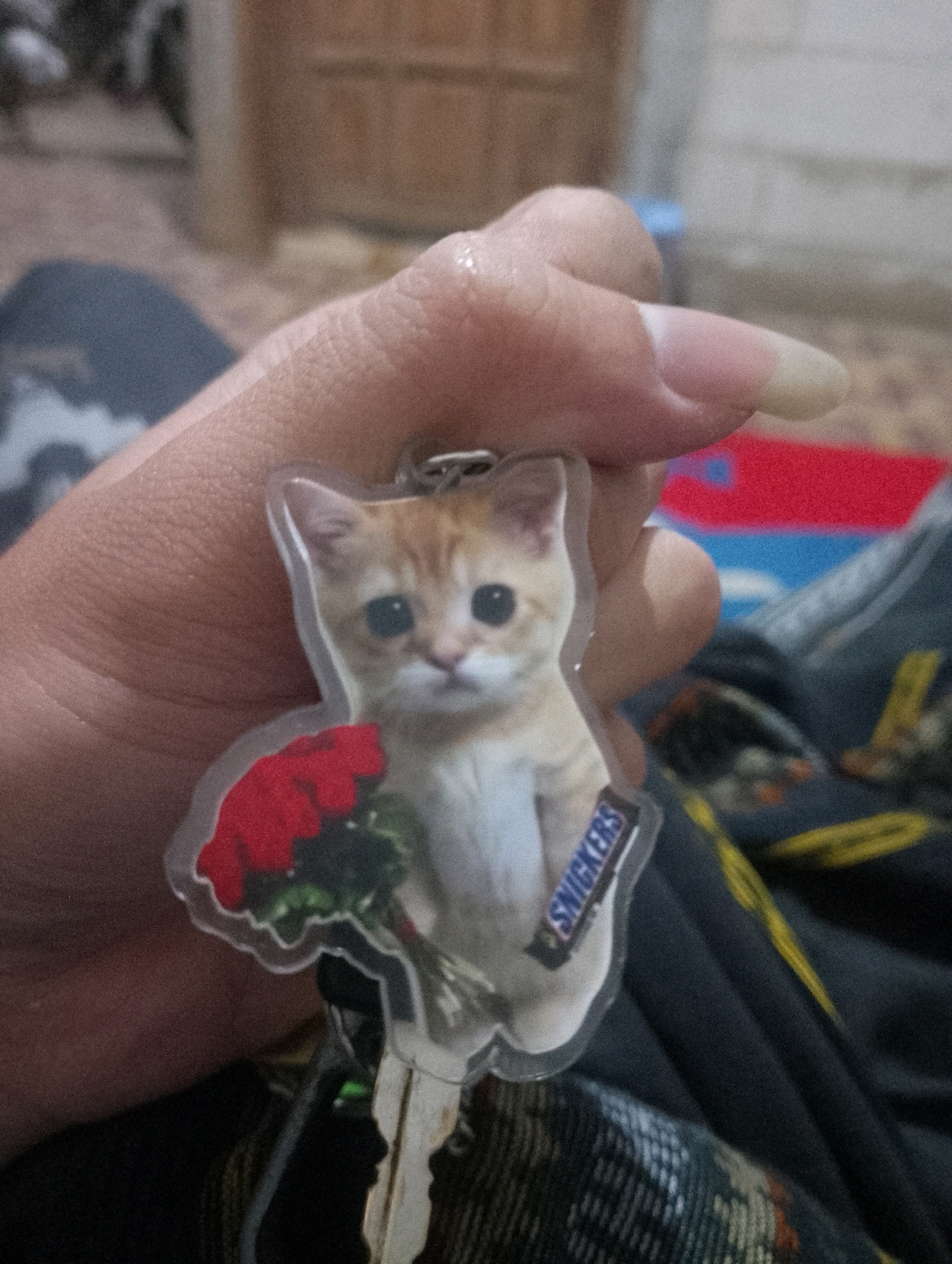 A close-up shot shows a hand holding a keychain with a clear plastic charm. The charm features a light orange tabby kitten with large, dark eyes, holding a bouquet of red roses. The kitten's right shoulder has a small "SNICKERS" logo. The hand has fair skin and a long, light-colored fingernail on the index finger. The background is blurred, showing what appears to be a patterned fabric and a wooden surface. The image was captured in Sowan Kidul, Indonesia.
