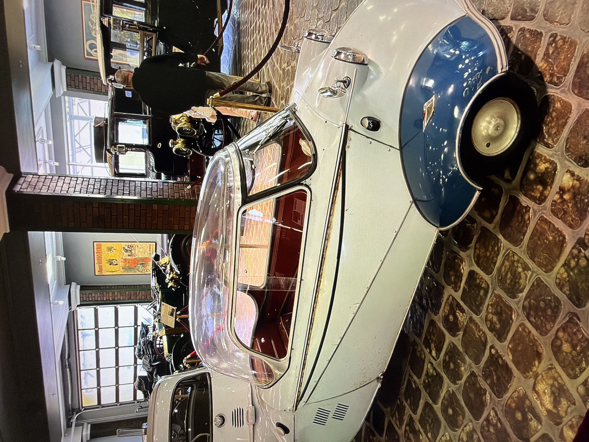 This image, captured in the city of Kotelniki, Russia, depicts a museum exhibit featuring vintage automobiles. The foreground is dominated by a white and blue three-wheeled microcar with a transparent bubble canopy. The car has a single wheel at the front and two at the rear, with the front wheel partially obscured by a blue fender. The interior of the car is visible, revealing red seating. The floor of the exhibit space is paved with a pattern of brown and grey rectangular stones.
In the background, several other classic cars are on display. To the left, partially visible, is another white car. Further back, a dark-colored vehicle with a tall, boxy design and brass accents is present, with a person in a dark jacket and grey pants walking away from the camera near it. Behind this, a vintage automobile with exposed engine components is showcased, with a poster featuring two figures and text in an unknown language displayed on the brick wall behind it. The overall environment suggests a well-lit museum or exhibition hall.
