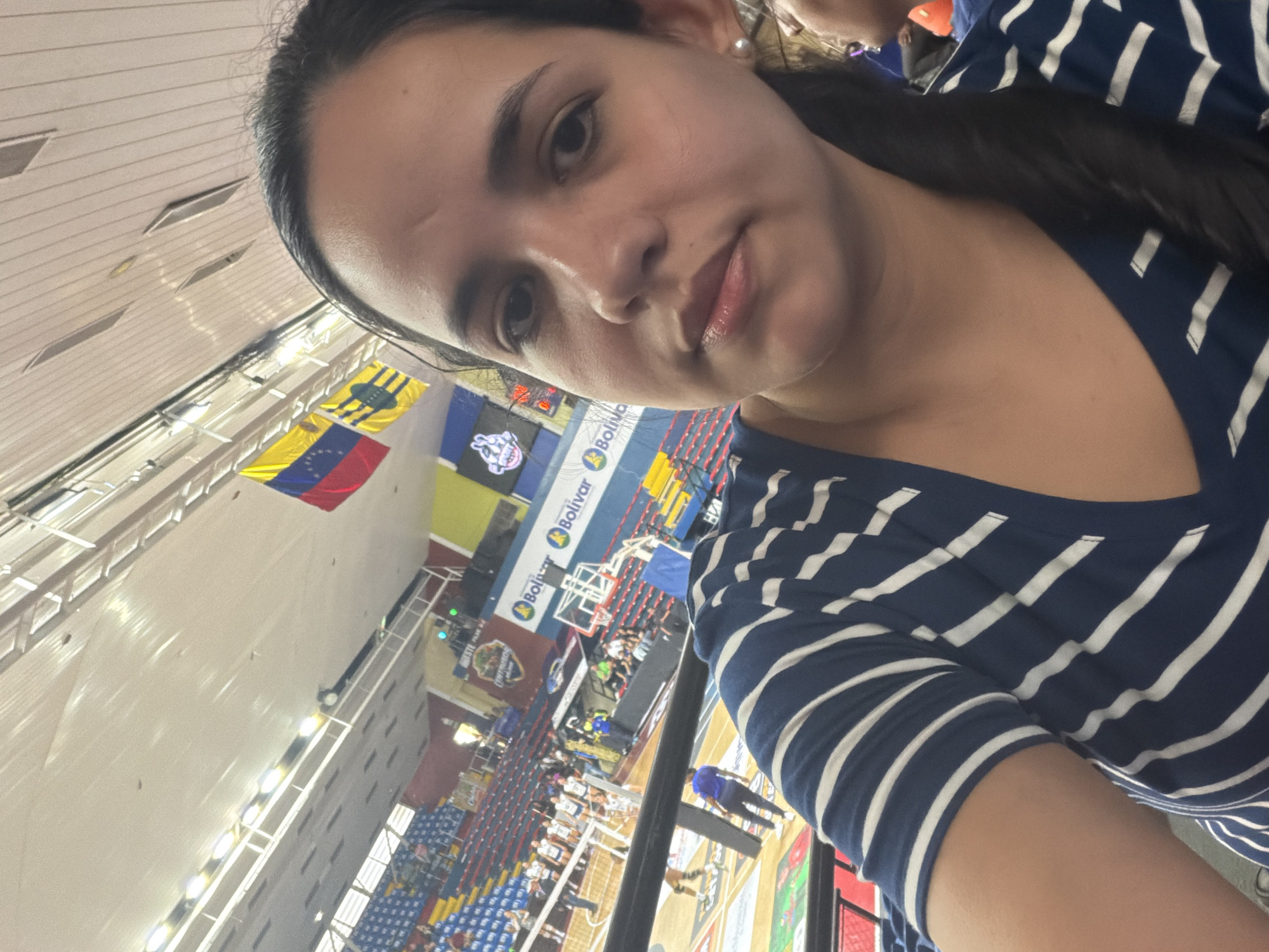 A woman is taking a selfie at a sporting event in Ciudad Guayana, Venezuela. The Venezuelan flag is visible in the background, along with banners with the text "Bolívar". The arena has red and blue seating. The woman is wearing a blue and white striped shirt. Other people are present in the arena, both in the stands and on the playing surface. The ceiling of the arena is visible, with linear lighting fixtures.