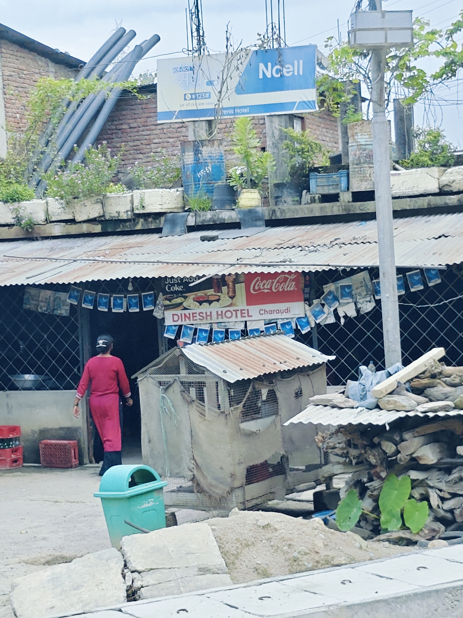 The image shows a street scene in Galchhi, Nepal. A woman in a red outfit walks away from the camera towards a building with a corrugated metal roof. Above the entrance to the building, a sign reads "Dinesh Hotel Charaudi" and a Coca-Cola advertisement is visible. Several flags with a blue crescent moon logo are strung across the front of the building. To the left of the entrance, there are red plastic crates stacked up. A green trash can is positioned on the ground in the foreground. In the background, a blue sign with "Ncell" is mounted on a pole. Various construction materials, including pipes and stones, are visible around the buildings.
