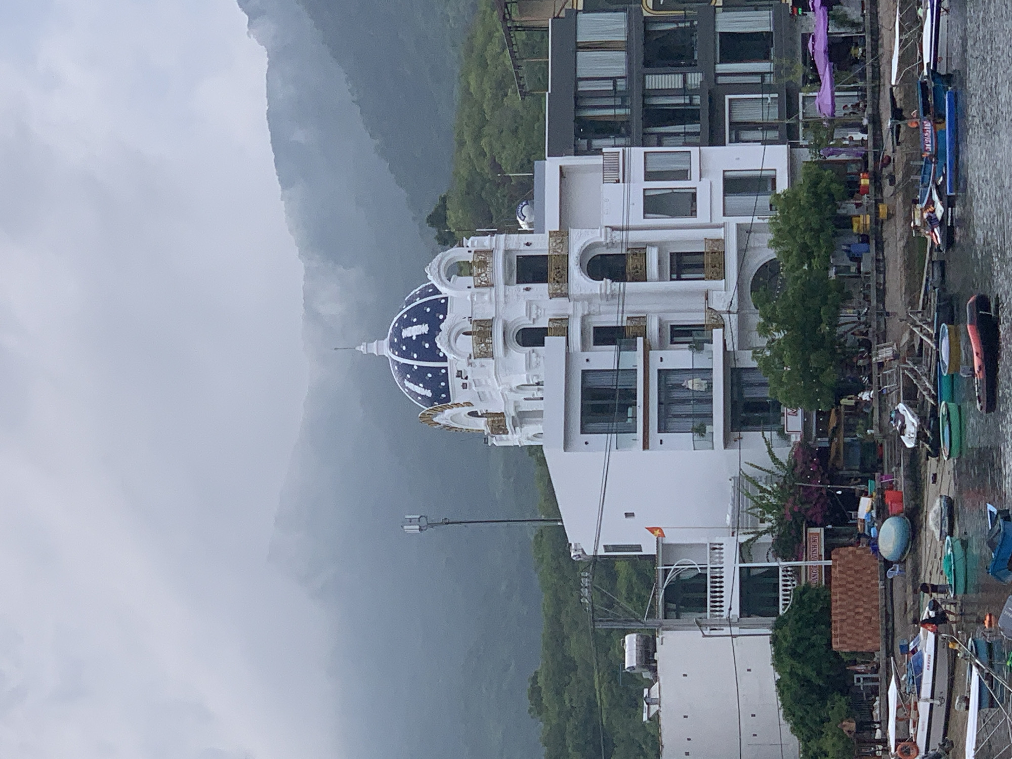 The image shows a waterfront scene in Vĩnh Hải, Vietnam, with a focus on a white building featuring a distinctive blue and white domed roof with gold accents. The building has multiple stories with numerous windows and balconies. To the right of the building, several boats are docked in the water, some appearing to be small fishing or tour boats. The water is calm, reflecting the overcast sky.
In the background, lush green mountains rise, partially obscured by mist or clouds, suggesting a humid or rainy climate. The weather appears to be overcast and possibly damp, as indicated by the muted lighting and the slightly hazy atmosphere.
The scene includes some people visible on the dock and possibly on the boats, but they are not the primary focus. There are also various objects like containers, umbrellas, and signage on the dock. One sign is partially visible with Vietnamese text that appears to read "VỊNH HẢI MINH BAY". The overall impression is a somewhat serene, albeit cloudy, day in a coastal or riverside town. The architectural style of the main building, with its dome and ornate details, stands out against the natural backdrop of the mountains.
