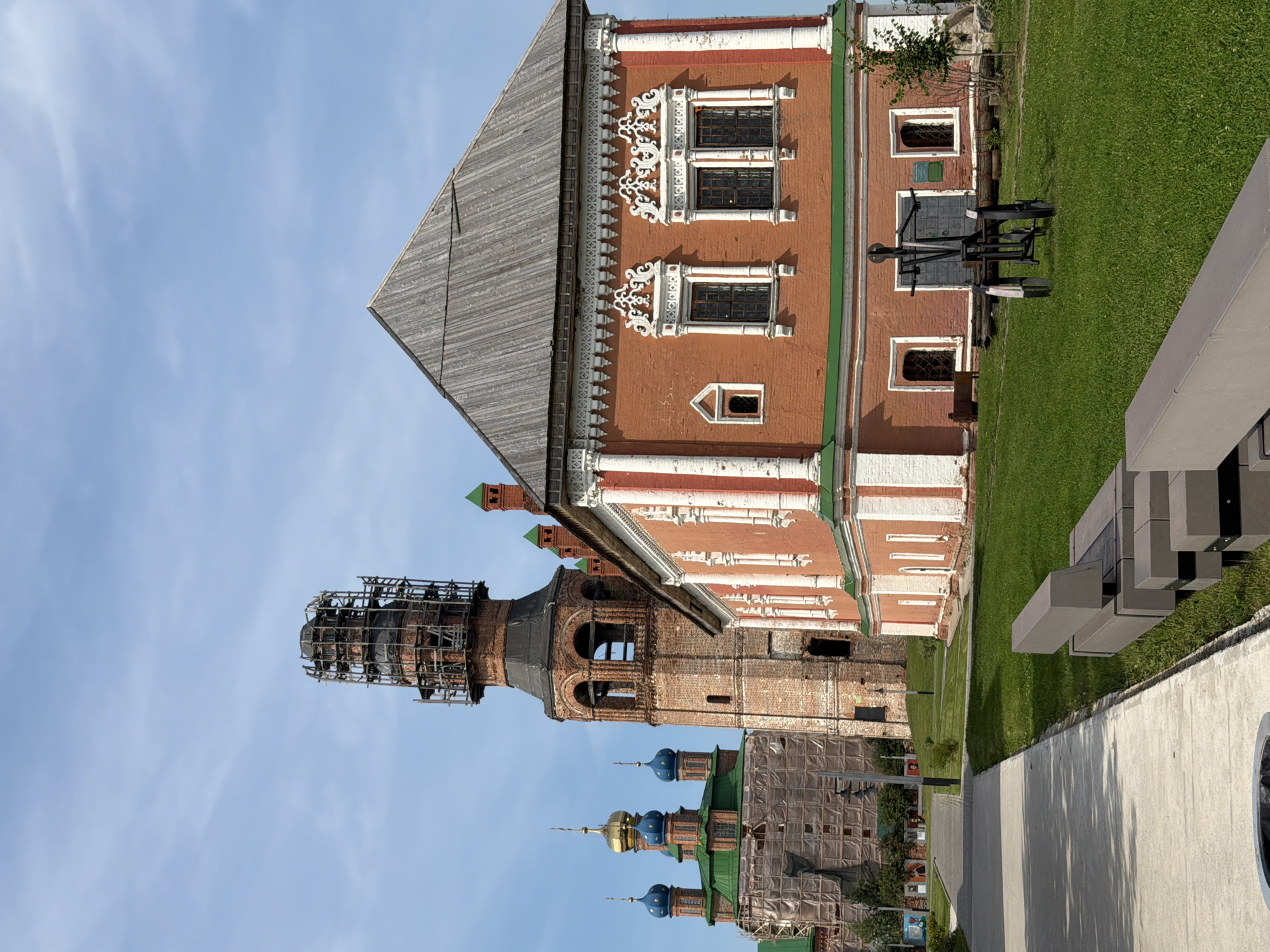 The image displays the Solovetsky Monastery in the city of Usolye, Russia, under a clear blue sky. In the foreground, a paved walkway runs along the left side of the frame, bordered by a well-maintained green lawn. A modern, concrete bench is visible on the grass to the right.
Dominating the mid-ground is a red brick building with white decorative trim around its windows and along the roofline. This building features a wooden, gabled roof. To the left of this structure, a tall, brick bell tower stands, partially covered in scaffolding. Behind the bell tower and to the far left, a complex of other monastery buildings is visible. Some of these structures have green roofs and golden or blue domes. One of these buildings is partially covered in scaffolding and what appears to be protective sheeting.
In the center of the lawn in front of the red brick building is a dark metal object on wheels, resembling a small, antique piece of artillery. Several informational signs and wooden logs are also visible near this object and the building. The overall scene suggests a historical site with ongoing restoration or maintenance work. There are no people visible in the image.