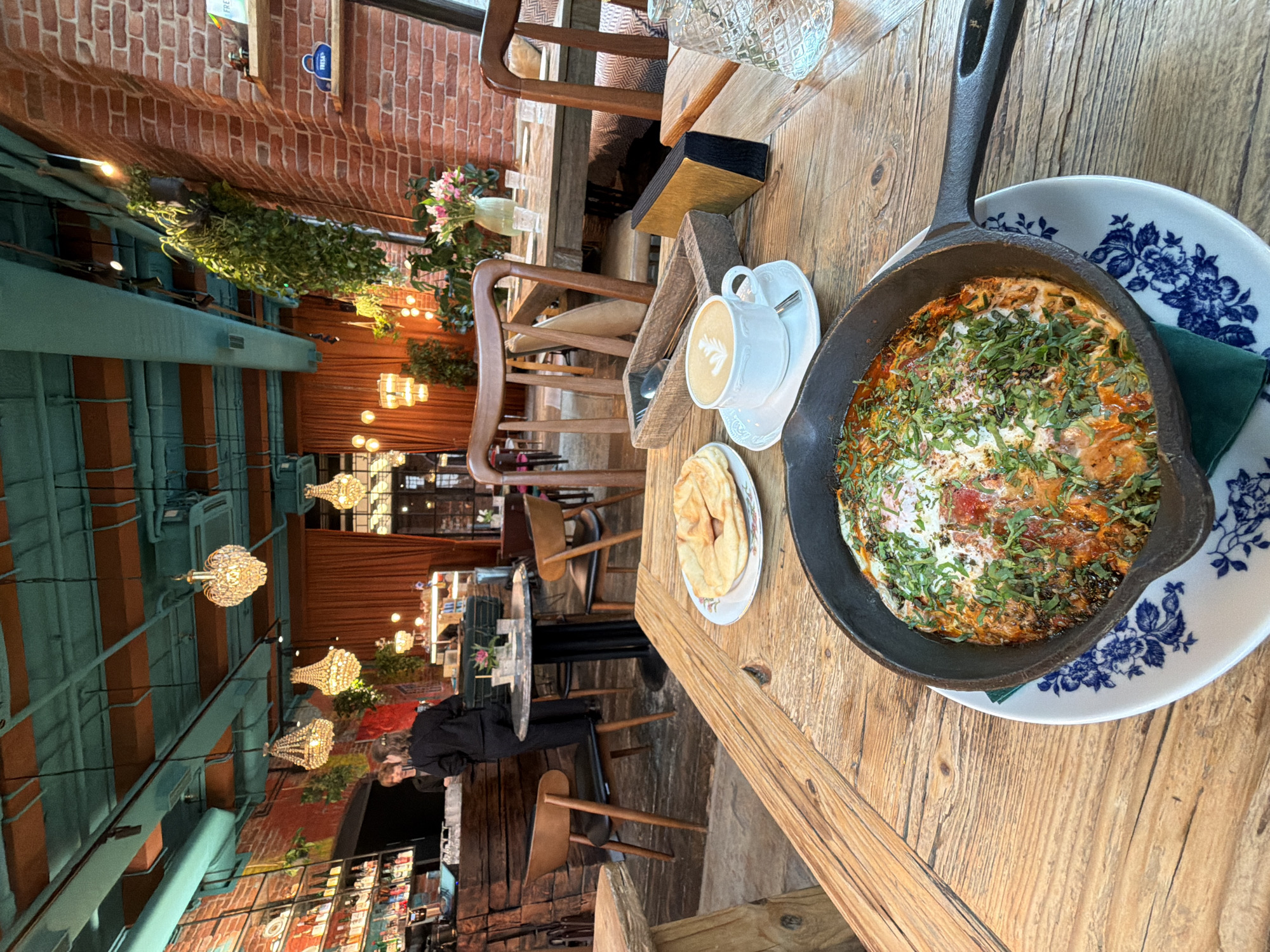 A first-person perspective shot shows a wooden table in a restaurant with a rustic design. In the foreground, a cast-iron skillet holds a dish topped with herbs and spices, served on a blue and white patterned plate. To the left, a small stack of flatbreads sits on a decorative plate. A white cup of coffee with latte art is positioned to the right of the flatbreads, alongside a wooden cutlery holder. In the background, other tables and chairs are visible, along with a bar area. The restaurant's interior features exposed brick walls, hanging plants, and decorative chandeliers. The scene is set in the city of Saint Petersburg, Russia.