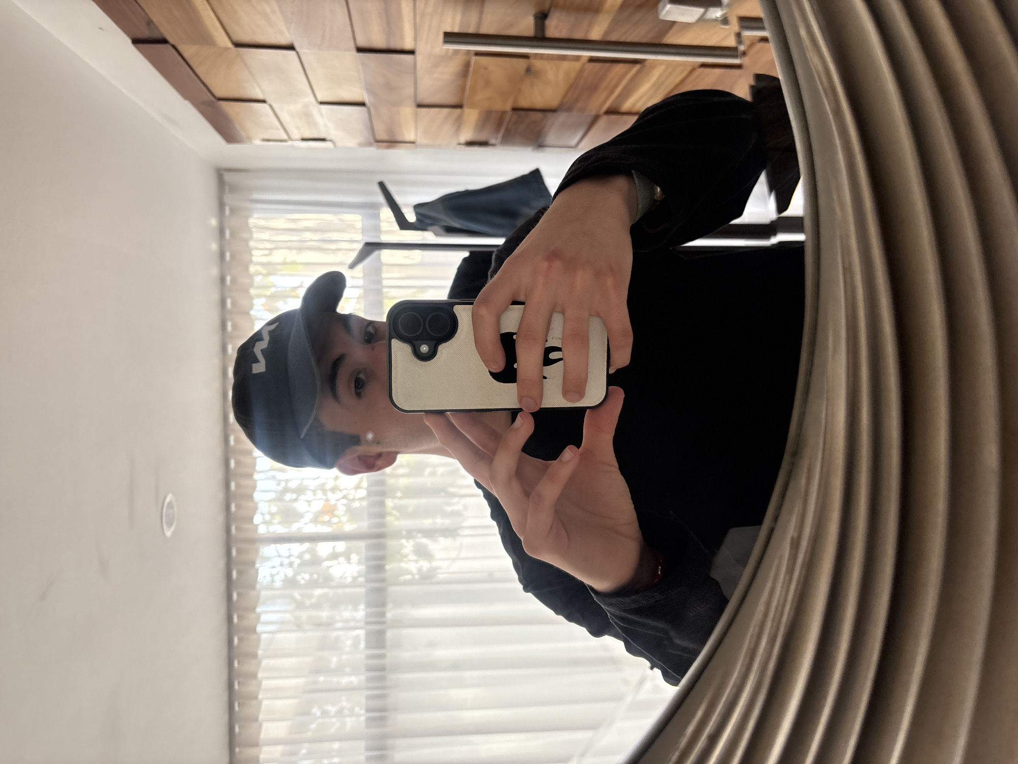 A person is taking a selfie in a mirror. The person is wearing a black baseball cap and a black jacket. The phone case is light with a black design on it. The mirror is round and has a rippled frame. Behind the person, a shelf with a dark item on it is visible, as well as a window with white curtains. The ceiling is wooden. The location appears to be indoors, but there is no specific information to confirm that this photo was taken in San Bernardino Tlaxcalancingo, Mexico.