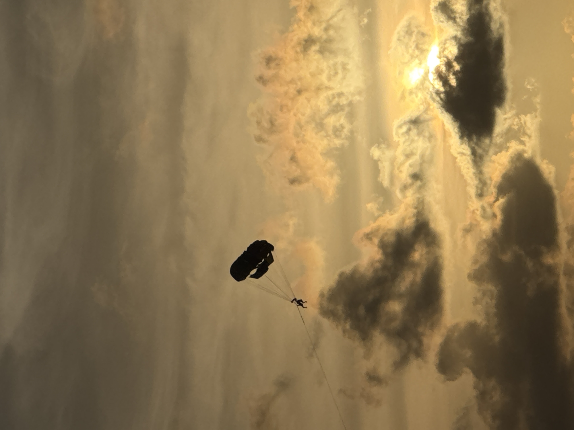An individual is parasailing across a hazy, sunset sky above Karon Beach in Karon, Thailand. The person, a dark silhouette, is suspended from a large, black parachute. The parachute is being pulled by an unseen boat. The sky is filled with clouds, some dark and ominous, others illuminated by the setting sun with a warm, golden hue. The overall scene suggests a recreational activity taking place in a tropical coastal environment.