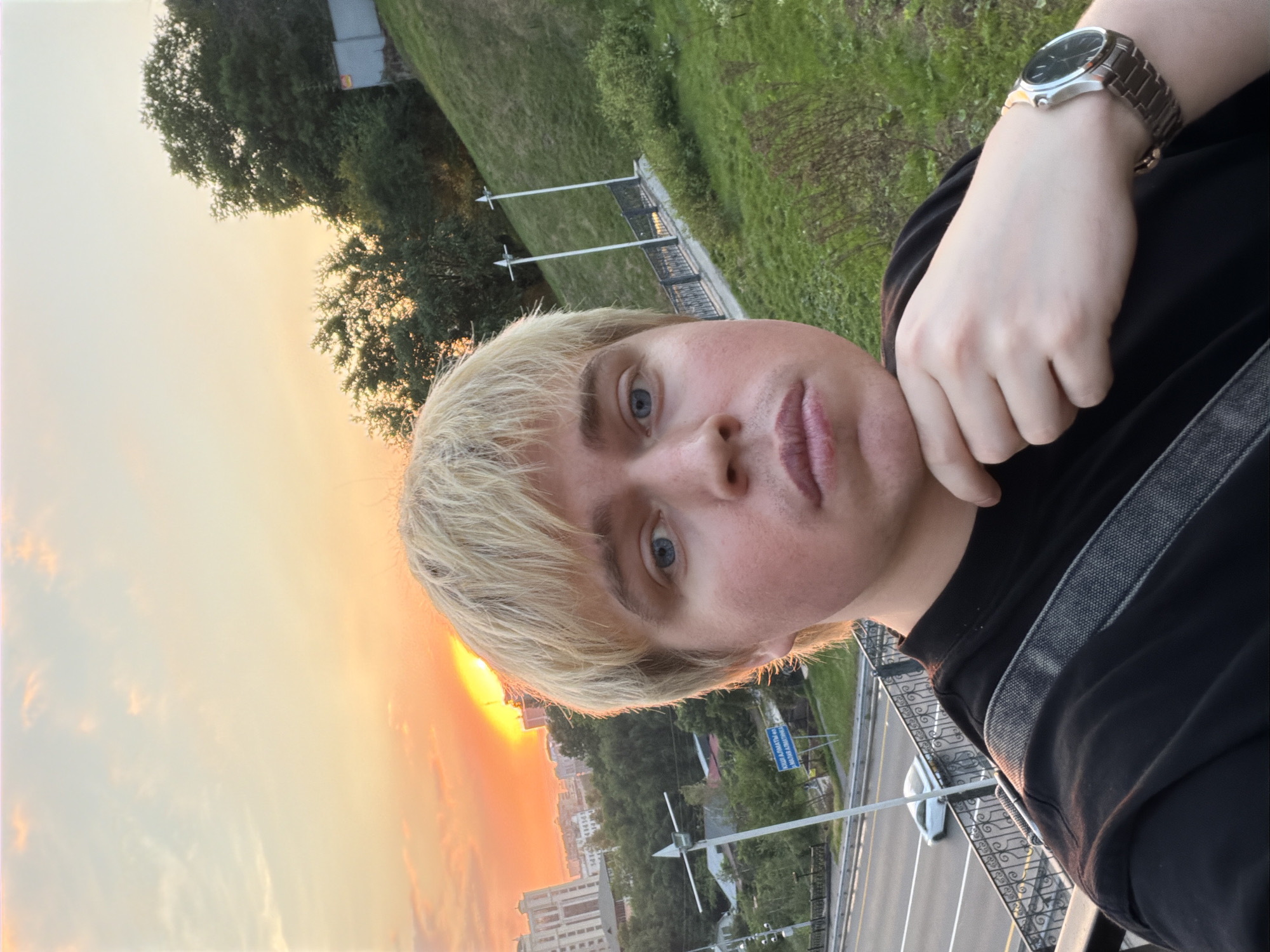 A person with blond hair and blue eyes is taking a selfie. They are wearing a black shirt and a silver watch. The background shows a sunset over a cityscape in Almaty, Kazakhstan. A highway with a car is visible, as well as green vegetation and buildings.