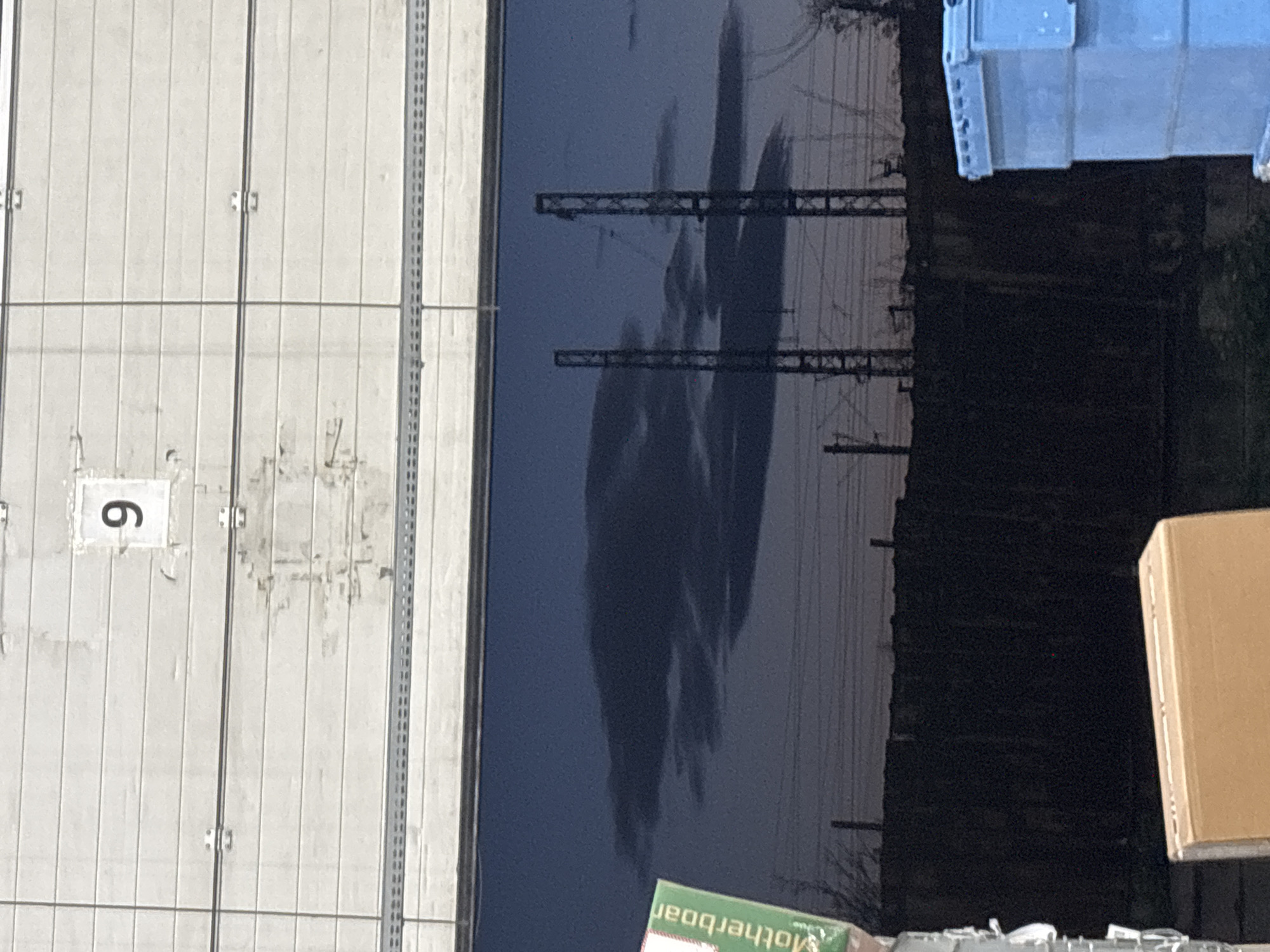 This image captures a static, industrial scene, likely near a railway or utility area, under daylight conditions. The composition is divided vertically.
On the left side of the frame, there is a large, light grey concrete panel wall, characterized by prominent vertical seams and smaller horizontal lines, indicating multiple panels or architectural details. Towards the upper left, a white square with the number "6" (appearing as a "9" if the image is viewed in its original upright orientation) is painted on one of these panels. There are also vertical metal support structures or railings attached to or alongside the concrete wall.
The right side of the image features a large, dark, highly reflective surface, possibly a very still body of dark water or a polished, dark ground surface. This surface acts as a mirror, reflecting a cloudy sky and a series of dark, intricate structures that appear to be overhead power lines or railway catenary lines against the lighter sky. The reflections are somewhat elongated and distorted, suggesting movement in the reflective medium or an angle of view.
In the foreground, closer to the viewer, several objects are visible. A light blue plastic container or bin is positioned at the top right. Below it, a light brown cardboard box is partially visible. Further down, towards the bottom right, a green box is seen with the word "Motherboard" printed on it, appearing upside down and reversed. Some scattered white debris is also present on the ground in this area.
The overall impression is one of an industrial or logistical setting, with man-made structures contrasting with natural light effects from the sky reflection. The time of day appears to be daytime, possibly late afternoon, given the atmospheric light. No human activity or specific event is taking place.