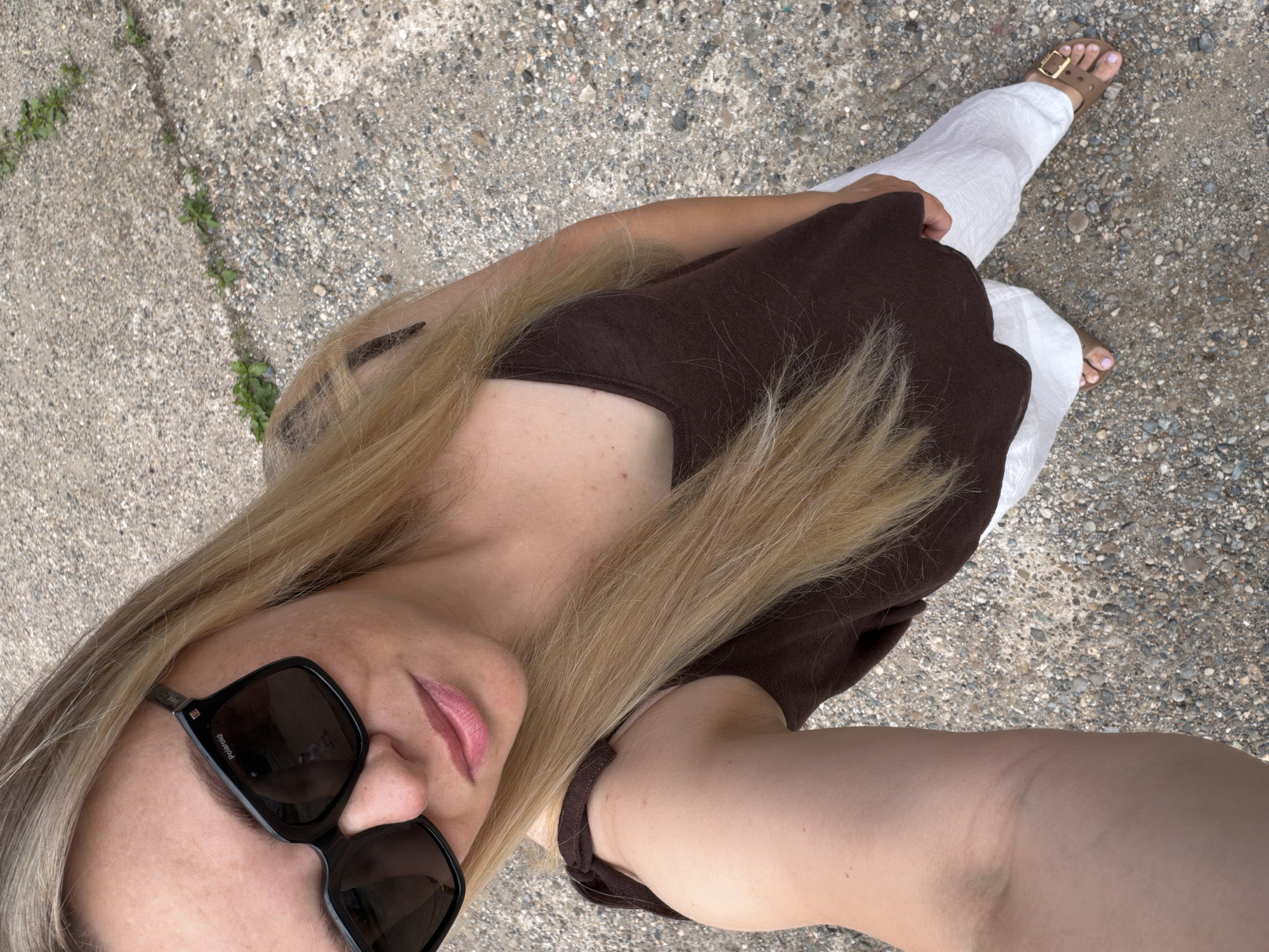 A blonde woman is taking a selfie outdoors, standing on a gravel path with patches of grass. She is wearing large black sunglasses with "Polaroid" written on the frame, a dark brown tank top, white linen pants, and brown sandals. Her right arm is extended, holding the camera. The ground is a mixture of concrete and small stones, with green plants growing in cracks. The scene appears to be in a public outdoor area, possibly a park or sidewalk in Votkinsk, Russia, based on the provided location.