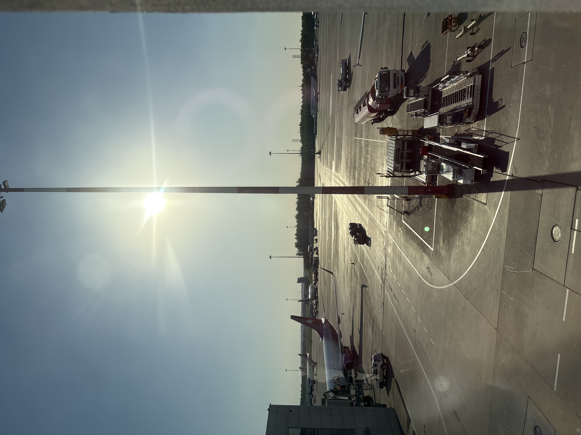 The image shows an airport tarmac with several aircraft, ground vehicles, and equipment. The scene is bathed in sunlight. A passenger jet with red tail is being serviced by ground crew. Other aircraft are parked nearby. The environment suggests the location is Pulkovo Airport in Saint Petersburg, Russia.