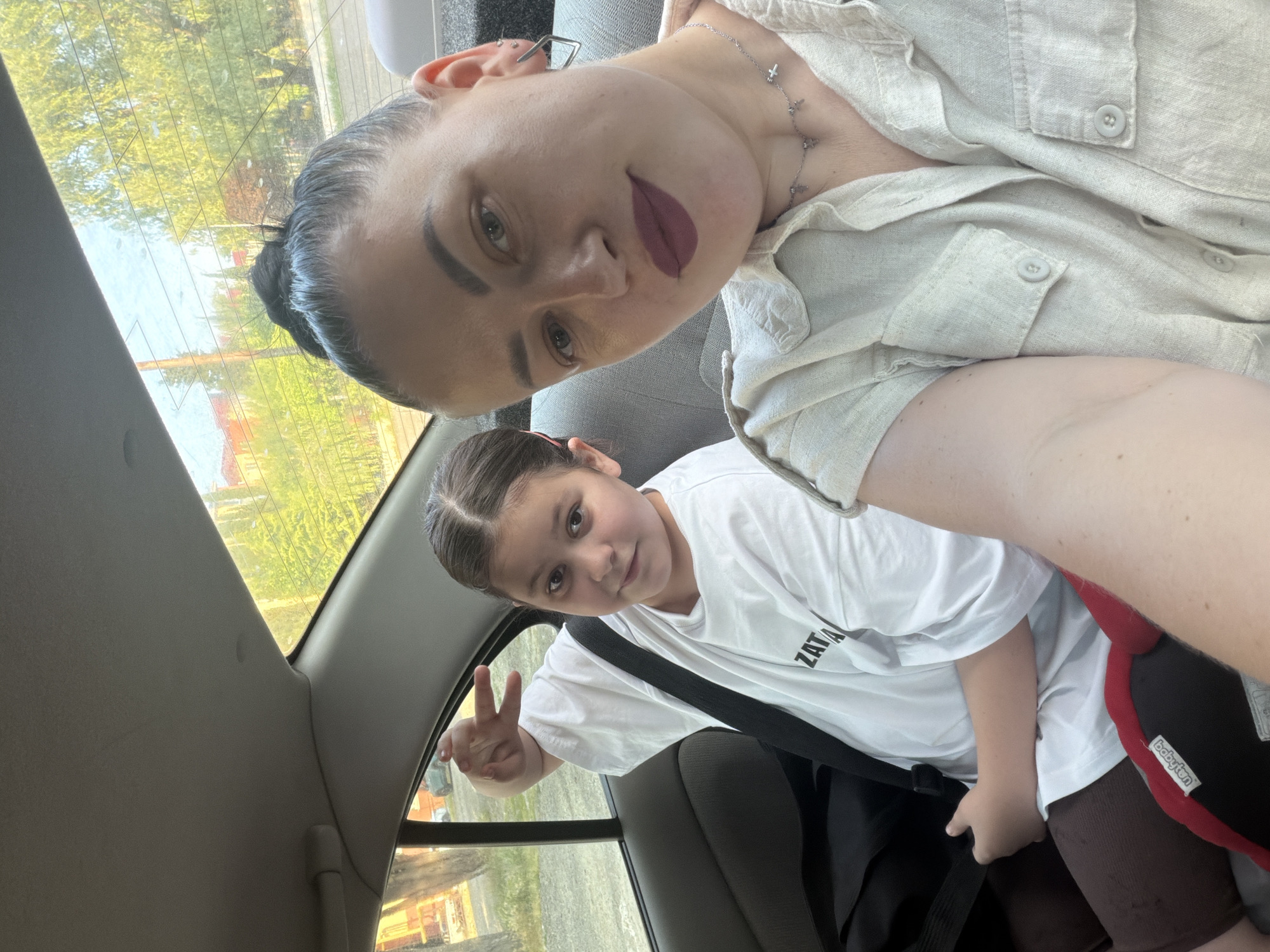 In Tuapse, Russia, a woman and a young girl are in the back seat of a car. The woman, on the right, is looking at the camera and wearing a light-colored, button-up shirt with two chest pockets and silver necklaces. Her hair is pulled back into a bun. The girl, on the left, is also looking at the camera and wearing a white t-shirt with some lettering on it. She has her seatbelt on and is holding up two fingers in a peace sign. The interior of the car is visible, including the grey seats and the car's headliner. Through the rear window, trees and buildings can be seen, suggesting a suburban or residential area. The lighting indicates it is daytime.