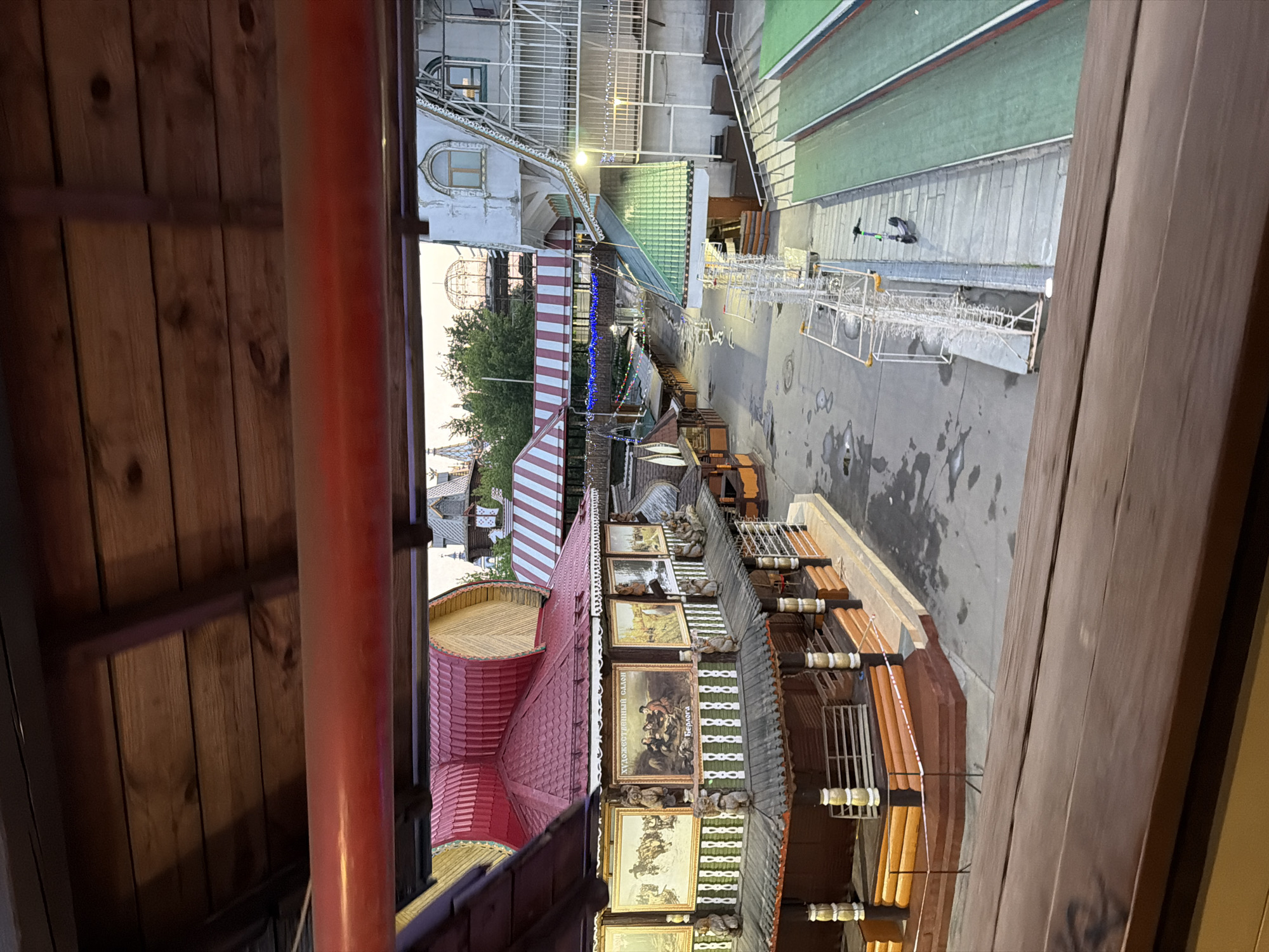View from an elevated position looking over a city landscape, presumably in Moscow, Russia, given the distinctive architecture. The red roofed structures and buildings with ornate decorations and paintings suggest a cultural or historical area. The presence of striped awnings, architectural details and construction scaffolding, imply the ongoing maintenance or restoration of these buildings. A public square or pedestrian zone is visible, possibly used for outdoor events or markets. The lighting indicates either dusk or early evening. No people are clearly visible in the photo.