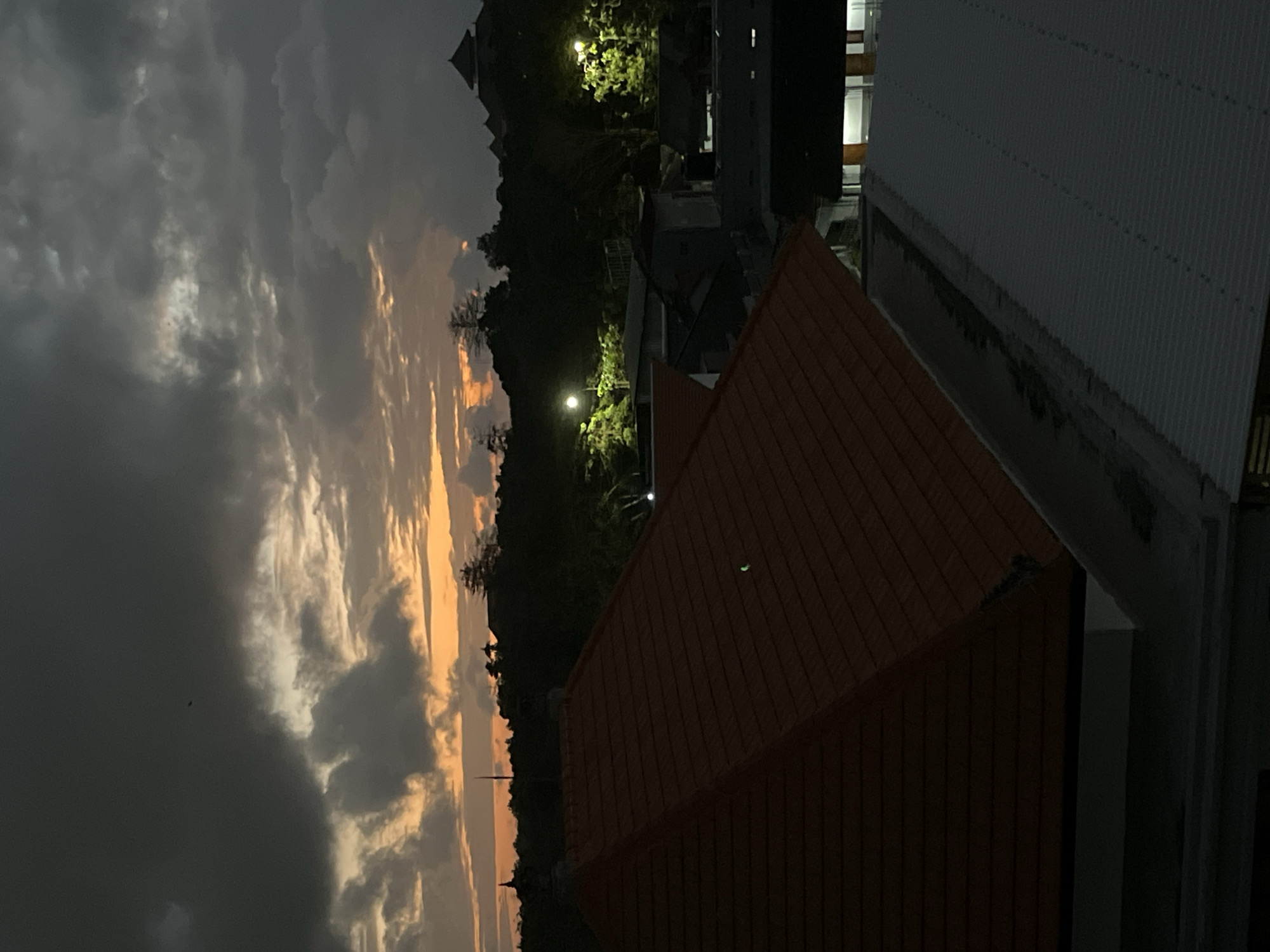 This media file shows an outdoor scene, likely at dusk or dawn, given the warm hues in the clouds and the artificial lighting visible in the distance. The composition is vertical, with the sky dominating the left side of the frame. The sky is filled with dramatic clouds, some dark and heavy, others tinged with orange and yellow light, suggesting the sun is low on the horizon or has recently set. A small dark silhouette, possibly a bird, is visible against the lighter clouds.
In the mid-ground, a dark, tree-covered hillside is visible, with lights illuminating some areas, hinting at human habitation or structures within the foliage. Silhouettes of buildings, including what appears to be a tower or spire, can be seen against the sky.
The foreground is occupied by the rooftops of buildings. The most prominent is a red-tiled roof with a triangular shape, suggesting a pitched roof. To the right of this, a lighter-colored, corrugated metal roof is visible, extending further into the background. The overall setting appears to be a developed area, possibly a town or resort, situated near a natural landscape with hills.
There are no people or specific activities clearly depicted. The mood is somewhat dramatic due to the sky's appearance, but also peaceful, conveying a sense of transition between day and night. The location context provided, Kuta Selatan, Indonesia, aligns with the tropical or sub-tropical vegetation that might be expected. No visible text is present in the image.