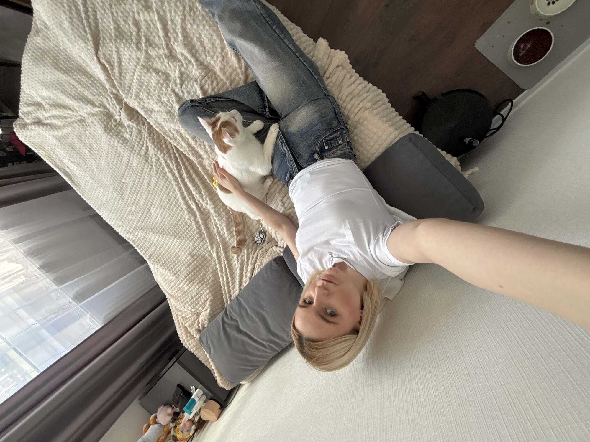 A high-angle shot depicts a blonde woman wearing a white t-shirt and blue jeans lying on a bed with a tan textured blanket. A white and orange cat is lying on her lap. Grey pillows are positioned behind her head and along her side. In the lower left, a window with grey curtains and a shelf with various small items is visible. To the right, on the floor, are a black bag and animal feeding dishes. The location cannot be confirmed as Vsevolozhsk, Russia, based solely on the image.
