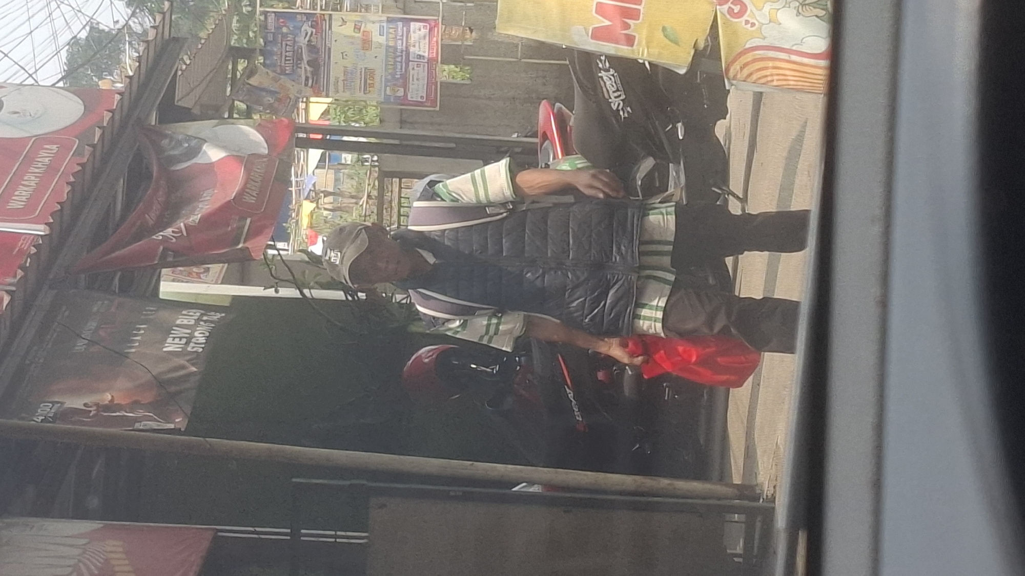 A person is walking outdoors in what appears to be a marketplace or busy street in Sirnagalih, Indonesia. The person is wearing a dark baseball cap, a black puffer vest over a horizontally striped t-shirt with green and white bands, and dark pants. They are carrying a red plastic bag in their left hand and a backpack on their shoulders. In the background, there are motorcycles parked, storefronts with banners and advertisements, and other people. The lighting suggests it is daytime, with sunlight casting shadows. The overall impression is a snapshot of everyday life in a bustling urban environment in Indonesia.