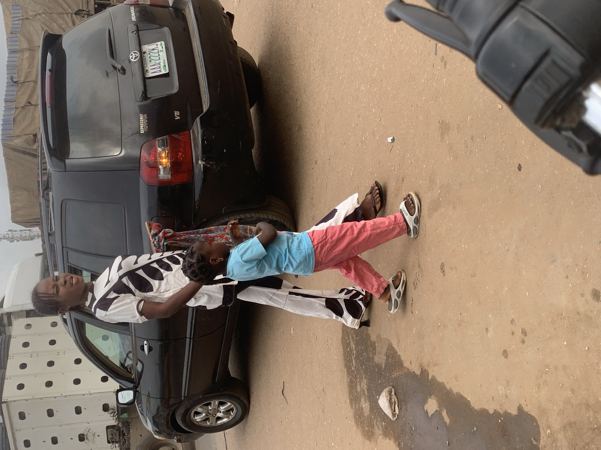 A young Black female, appearing to be in her late teens or early twenties, stands to the left of a black Toyota Highlander SUV, holding a bundle of fabric. She is wearing a black and white patterned t-shirt and black and white patterned pants. A younger Black female child, about 4-5 years old, walks in front of her to the right. The child is wearing a light blue t-shirt and pink pants, with white sandals. The SUV has a license plate that reads "AAA-222KJ". The scene appears to be on a dirt or unpaved road, with a large white truck visible to the left behind the SUV, and another large vehicle with a tarp covering it in the background. The ground is a light brown color, with some patches of wetness and scattered debris. The image was taken in Shagamu, Nigeria.