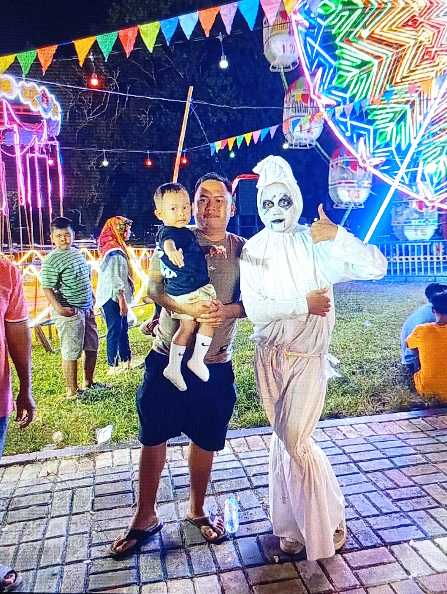 In Lhokseumawe, Indonesia, a man stands holding a young child. The man is wearing a dark shirt and black shorts, while the child is in a dark blue t-shirt and white socks. Beside them, a person in a white costume with face paint resembling a ghost poses with their thumb up. In the background, a brightly lit Ferris wheel and carnival rides, adorned with colorful flags and lights, create a festive atmosphere. Other people are visible in the vicinity, with some seated and others standing. The ground is paved with bricks, and grass is present in some areas, suggesting an outdoor event or fairground setting. The overall scene depicts a nighttime gathering at a carnival.