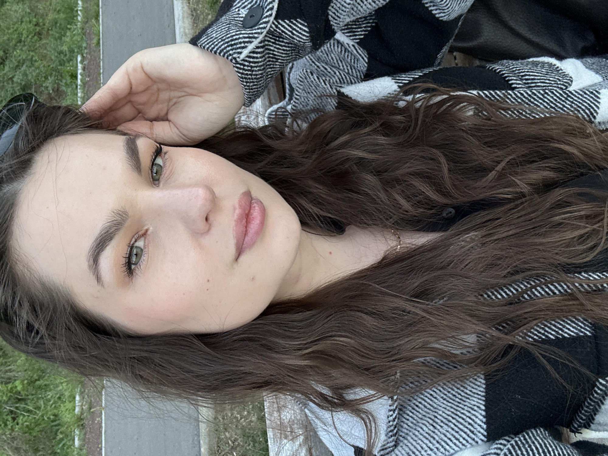 A young woman with brown curly hair and fair skin poses for a selfie. She has green eyes, full lips, and well-defined eyebrows. Her left hand is raised to her head. She is wearing a black and white plaid jacket. The background shows a glimpse of greenery and a paved surface, potentially a sidewalk or street. The location is Chaikovsky, Russia.
