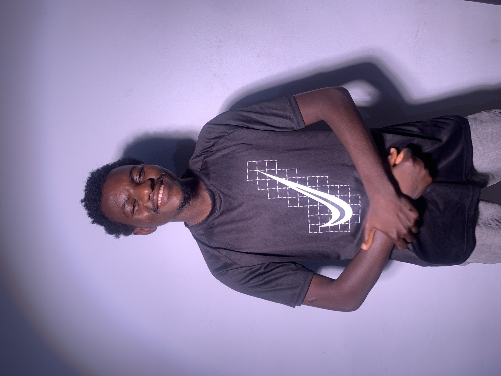 A young Black man with curly hair and a beard smiles broadly at the camera, tilting his head slightly to his right. He is wearing a dark t-shirt with a white graphic design on the chest, possibly a stylized logo, and grey shorts. His arms are crossed, with his hands clasped over his abdomen. The lighting in the image is dim, casting a shadow on the wall behind him. The background is a plain, light-colored wall, and the overall scene appears to be indoors, with no clear indications of time of day, weather, or specific location. The man's expression conveys a sense of happiness or amusement.