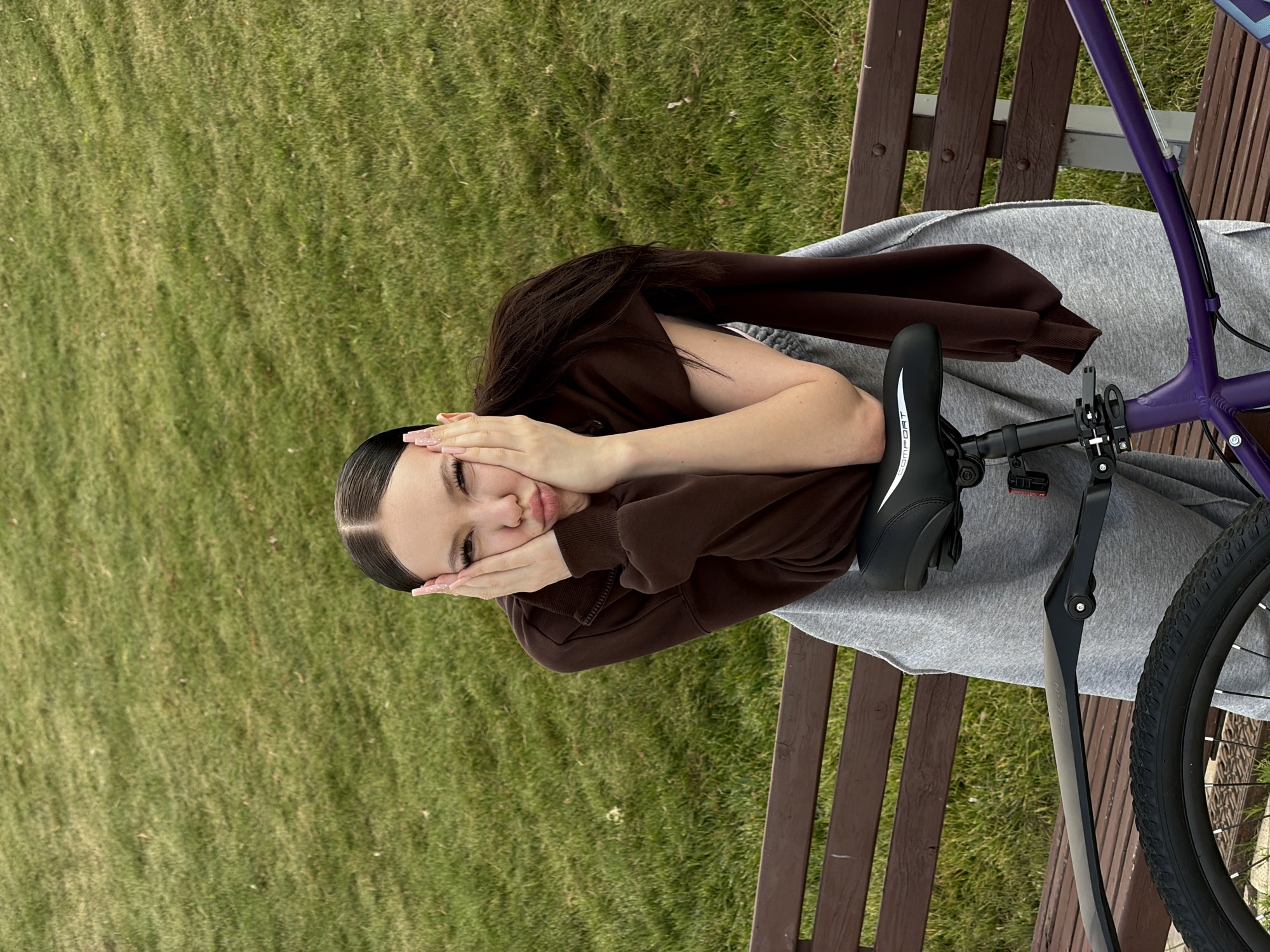 A young woman with dark hair pulled back into a ponytail sits on a park bench, leaning on the seat of a purple bicycle. She is wearing a brown zip-up hoodie over a grey outfit and has her hands pressed against her cheeks, with her lips pursed. The bicycle has a black seat and a black fender over the rear wheel. The bench is made of dark wood. The background consists of a grassy area, suggesting an outdoor park setting in Krasnogorsk, Russia.