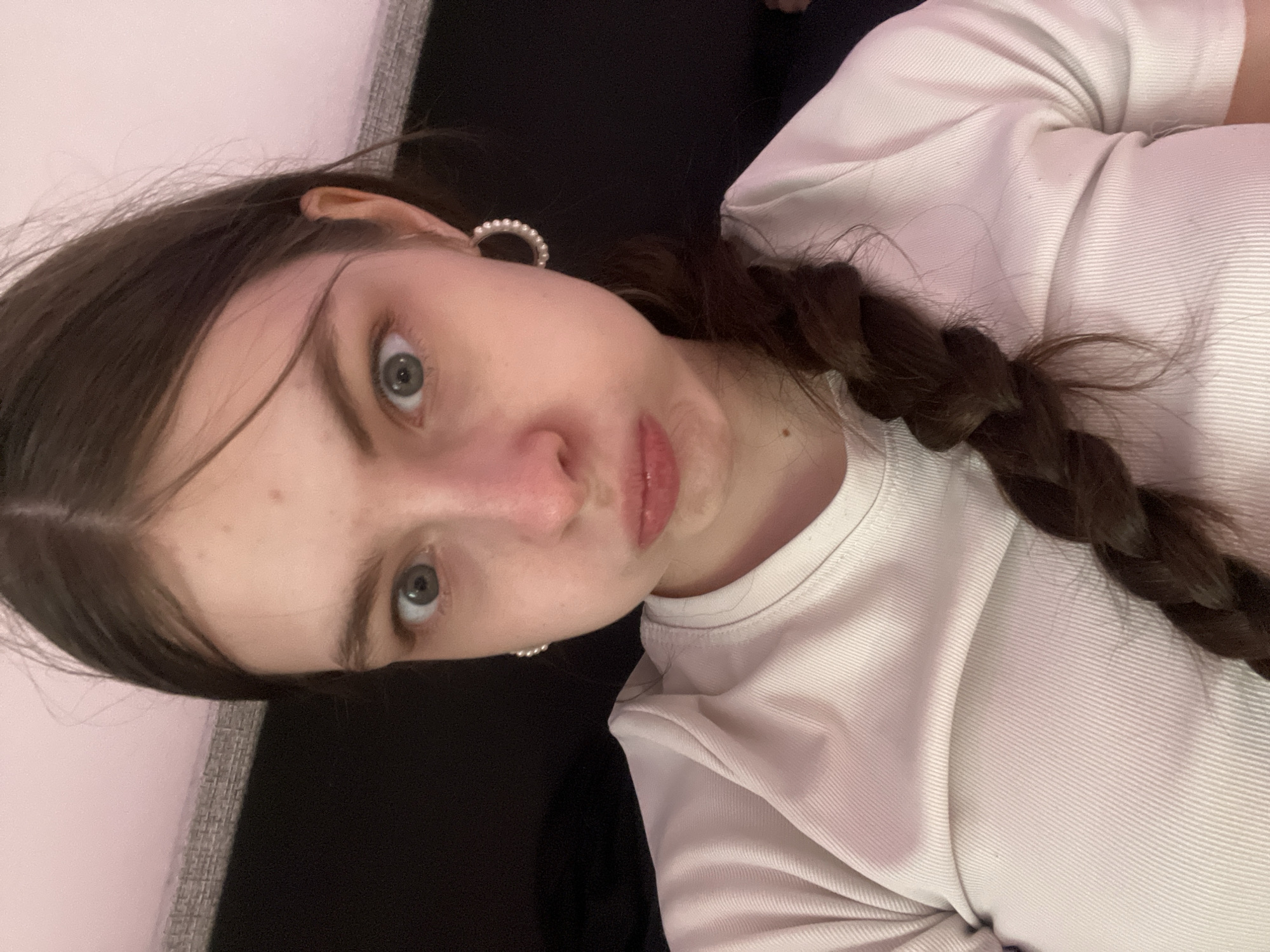 A young woman with light skin, gray-blue eyes, and light brown hair braided on the right side of her face is posing for a photo. She is wearing a white, ribbed T-shirt and pearl hoop earrings. She has a slight pout and is looking directly at the camera. The background consists of a light gray surface and a dark surface, possibly a couch. Without location specific data, it is not possible to determine if this image was taken in Saint Petersburg, Russia.