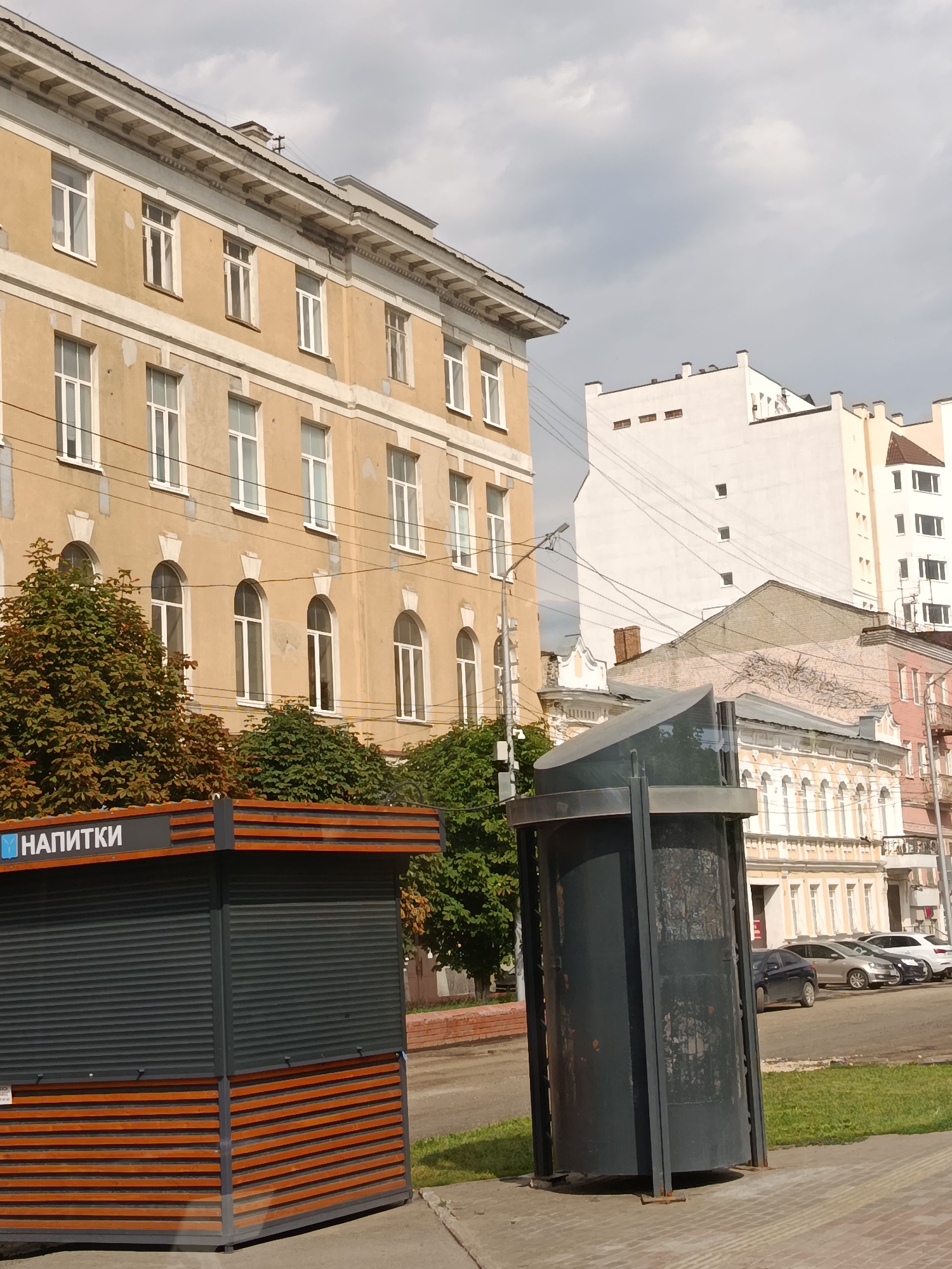 The image shows a street scene in Saratov, Russia, on a cloudy day. In the foreground, there is a modern kiosk with the word "НАПИТКИ" (drinks) visible on its side. Adjacent to the kiosk is a cylindrical, dark-colored trash receptacle with a futuristic design. Behind these structures, a multi-story building with a light yellow facade and white-framed windows dominates the left side of the frame. Further down the street, other buildings of varying architectural styles and heights are visible, including a white building with square windows and a cream-colored building with arched windows. Several cars are parked along the street. Lush green trees are present on the left side of the image, partially obscuring the lower floors of the yellow building. The sky is overcast with gray clouds.