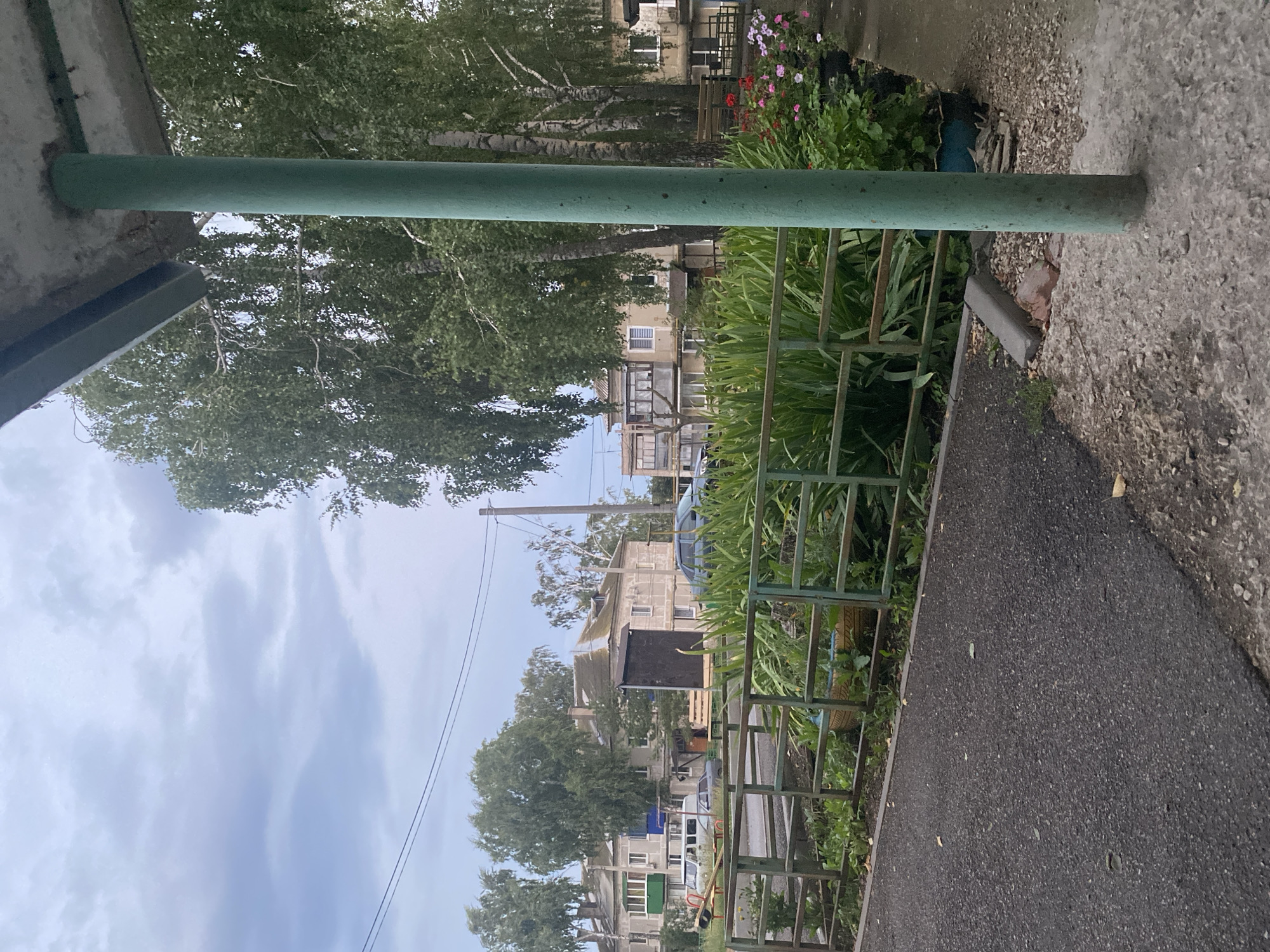 A vertical, eye-level, medium shot captures an outdoor scene on a cloudy day in Oktyabrsk, Russia. The foreground features a dark asphalt path on the left, bordered by a low, green metal fence. Beyond the fence, dense green foliage and a few potted plants with pink and red flowers are visible. A prominent, weathered green cylindrical pillar stands to the right of the frame, partially obscuring the background. In the mid-ground, apartment buildings with pitched roofs, some with balconies, are seen. Several cars are parked along the street in front of the buildings. Birch trees with white bark and green leaves are interspersed throughout the scene, with a few taller, more wispy trees on the left. The sky is overcast with soft grey clouds.