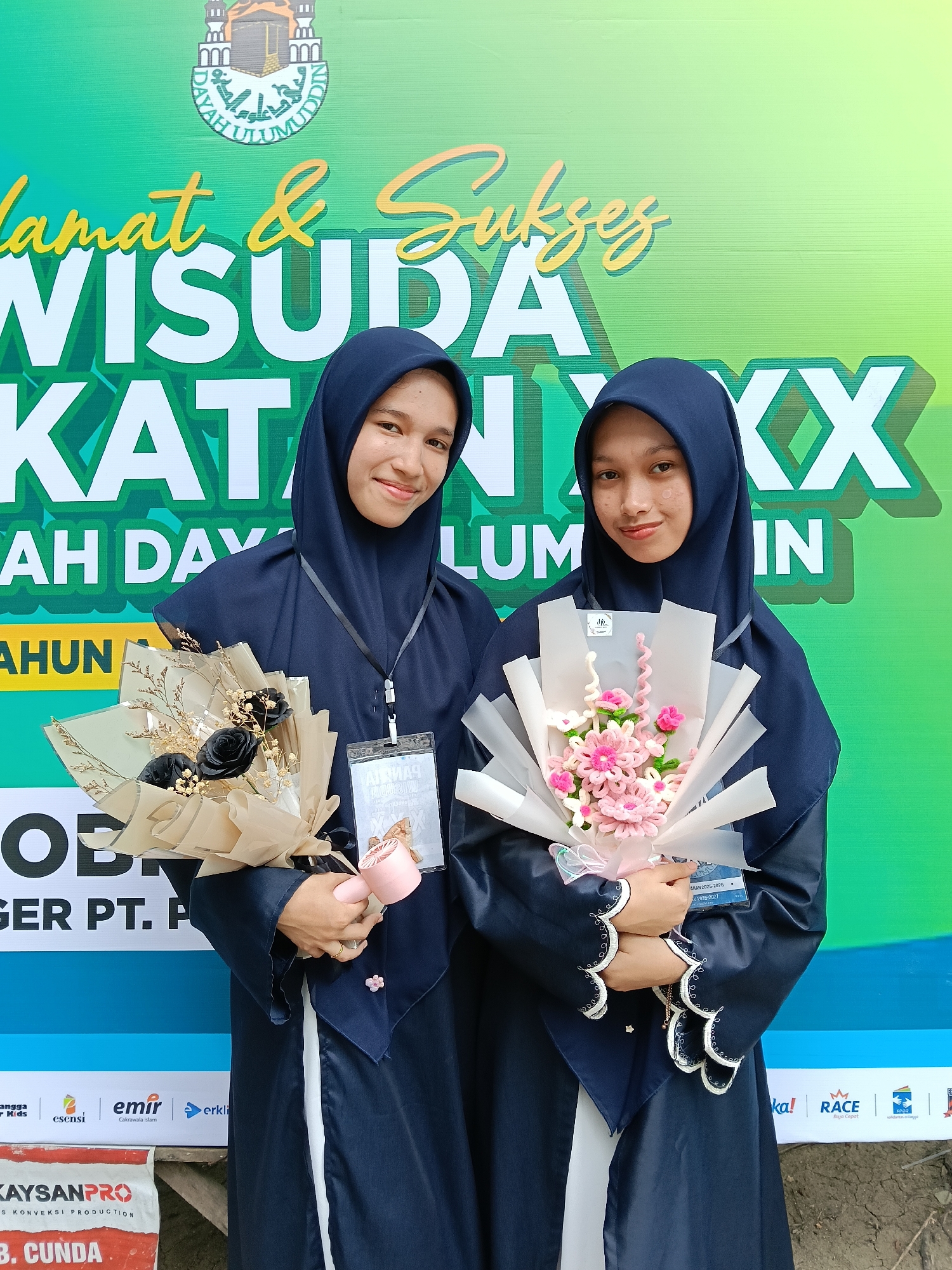 Two young women in dark blue hijabs and graduation gowns stand side-by-side in front of a green and yellow banner with the words "Selamat & Sukses WISUDA KATAN XXX" displayed on it. The banner also includes the logo of "DAYAH ULUMUDDIN" and mentions "TAHUN". The women are holding bouquets of flowers; one holds a bouquet with black roses and the other a bouquet with pink crocheted flowers. Each woman is wearing a lanyard with a badge. The banner appears to be part of a promotional setup with sponsor logos visible along the bottom, including "KAYSANPRO" and "emir", among others. The location is Lhokseumawe, Indonesia, as implied by the banner's content and logos. The scene appears to be a graduation celebration or photo opportunity.