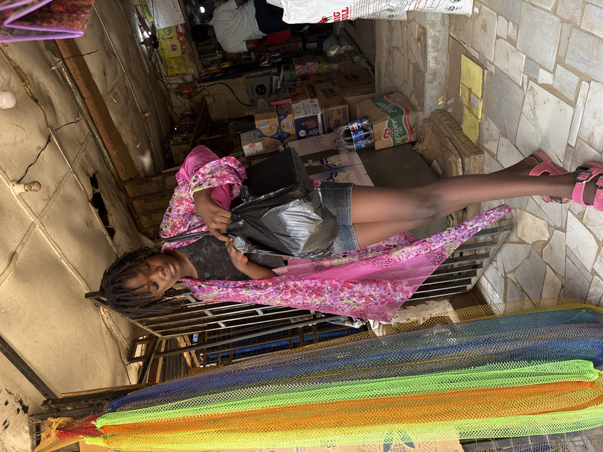 A young Black girl sits with her legs outstretched and slightly elevated, in what appears to be a small shop in Abuja, Nigeria. She is wearing a black top, denim shorts, pink sandals, and a pink floral shawl. She holds a black plastic bag. Behind her are stacks of colorful netting. In the background, the shop contains boxes of goods. Another person is visible in the background of the shop. The floor is tiled.