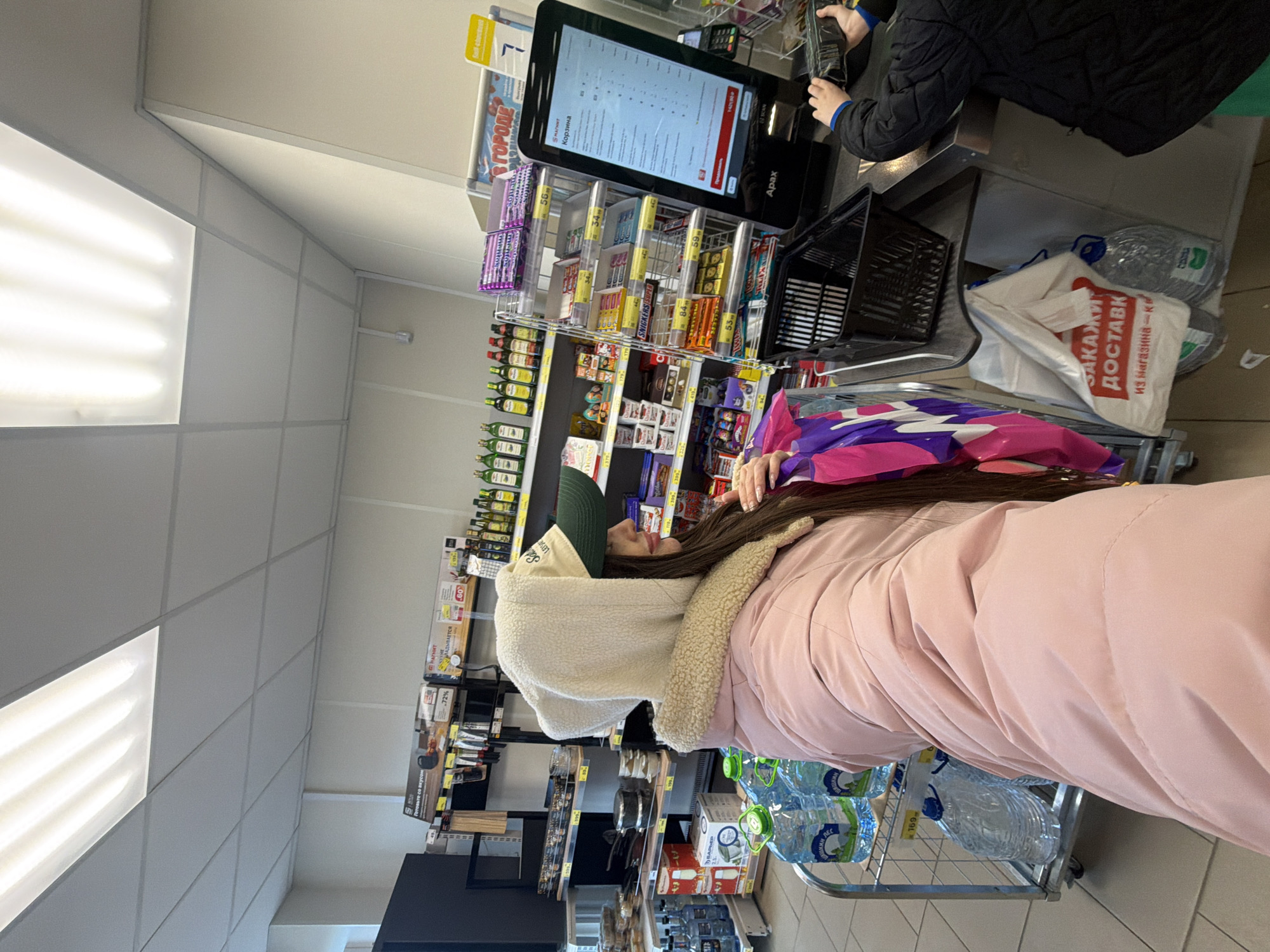 Here is a description of the image:
**Setting:**
The image appears to be taken inside a "Magnet" grocery store. The location is likely in Russia, possibly the city of Saint Petersburg, based on the Cyrillic script on signs, products, and a delivery bag indicating "from the store." The store has fluorescent lighting on the ceiling and shelves stocked with various goods.
**People:**
A woman wearing a green cap and a pink coat is standing with a shopping cart filled with bottled water. Another person is partially visible at a self-checkout kiosk, likely in the process of scanning and paying for their groceries.
**Objects:**
The store shelves are stocked with a variety of goods, including beverages, snacks, and wine. The shopping cart is holding multiple bottles of water. A self-checkout kiosk is visible with a screen displaying a "Корзина" (Basket) and total amount. A delivery bag with "ЗАКАЖИ ДОСТАВК из магазина" is visible next to the self-checkout.
**Actions:**
The woman is standing by her shopping cart. Another person seems to be operating the self-checkout kiosk.
**Interactions:**
The woman may be waiting in line for the self-checkout. The other person is actively using the self-checkout to purchase goods.
**Environment:**
The grocery store is orderly and well-lit.