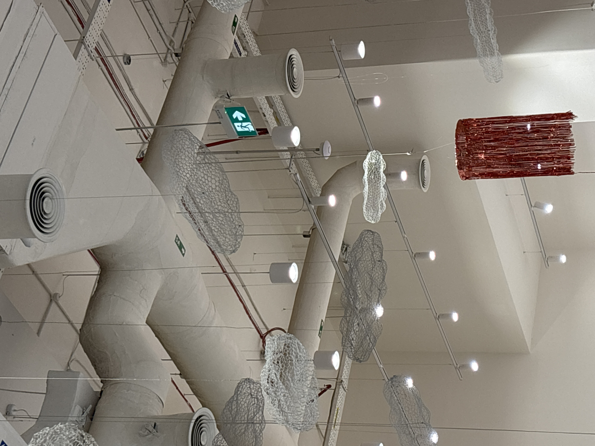 This image shows the interior ceiling of a public space, likely a mall or retail store, in Dubai, United Arab Emirates. The ceiling features exposed HVAC ductwork, some of which is covered with a rough, textured material. Suspended from the ceiling are several decorative elements resembling clouds, made of white, translucent mesh. There are also numerous track lights and recessed ceiling lights illuminating the area. A prominent green and white exit sign with a running figure is visible on one of the vertical ductwork sections. In the lower right portion of the image, a cylindrical red decorative fixture, possibly a chandelier or lamp with fringe, hangs. The overall impression is a modern, retail-inspired environment with artistic ceiling installations.