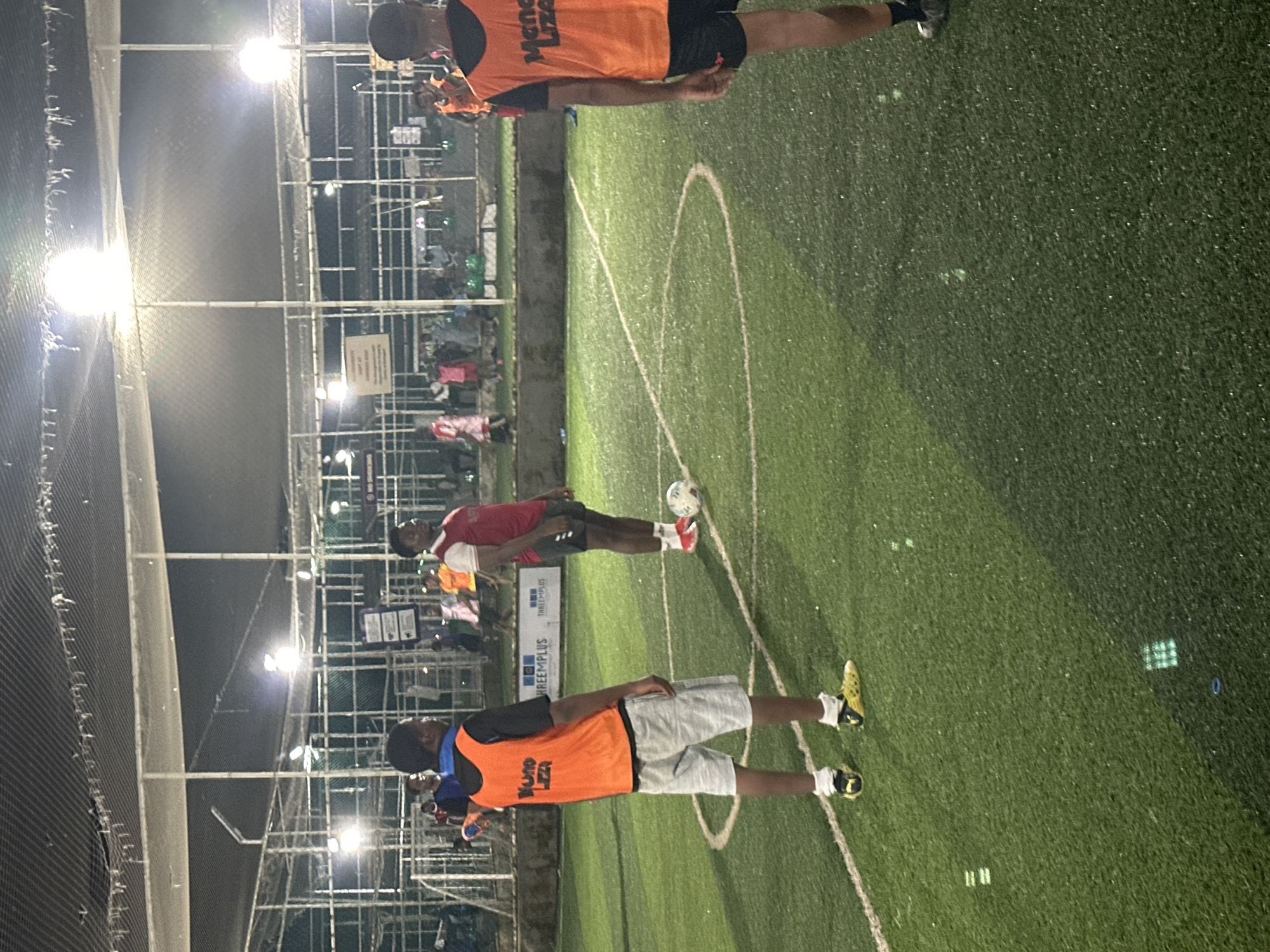 The image depicts an outdoor football pitch at night, illuminated by bright overhead lights. The pitch appears to be artificial turf and is marked with white lines, including a penalty box. Several individuals, presumably participating in a football match or practice, are present on the field. In the foreground, two young men are visible. The man on the left is wearing an orange vest over a black t-shirt and light gray shorts, with yellow and black football boots. He is facing away from the camera and appears to be standing. To his right, a second man, also wearing an orange vest, is standing near a football. He is wearing dark shorts and red and white football boots. In the background, more people are visible, some wearing similar vests, suggesting a team activity. The setting includes fencing around the pitch and structures that suggest a sports complex or recreational area. The overall atmosphere is one of an active sporting event at night. This scene is consistent with the type of recreational facilities found in urban areas like Abuja, Nigeria, which often feature illuminated sports grounds for evening use.
