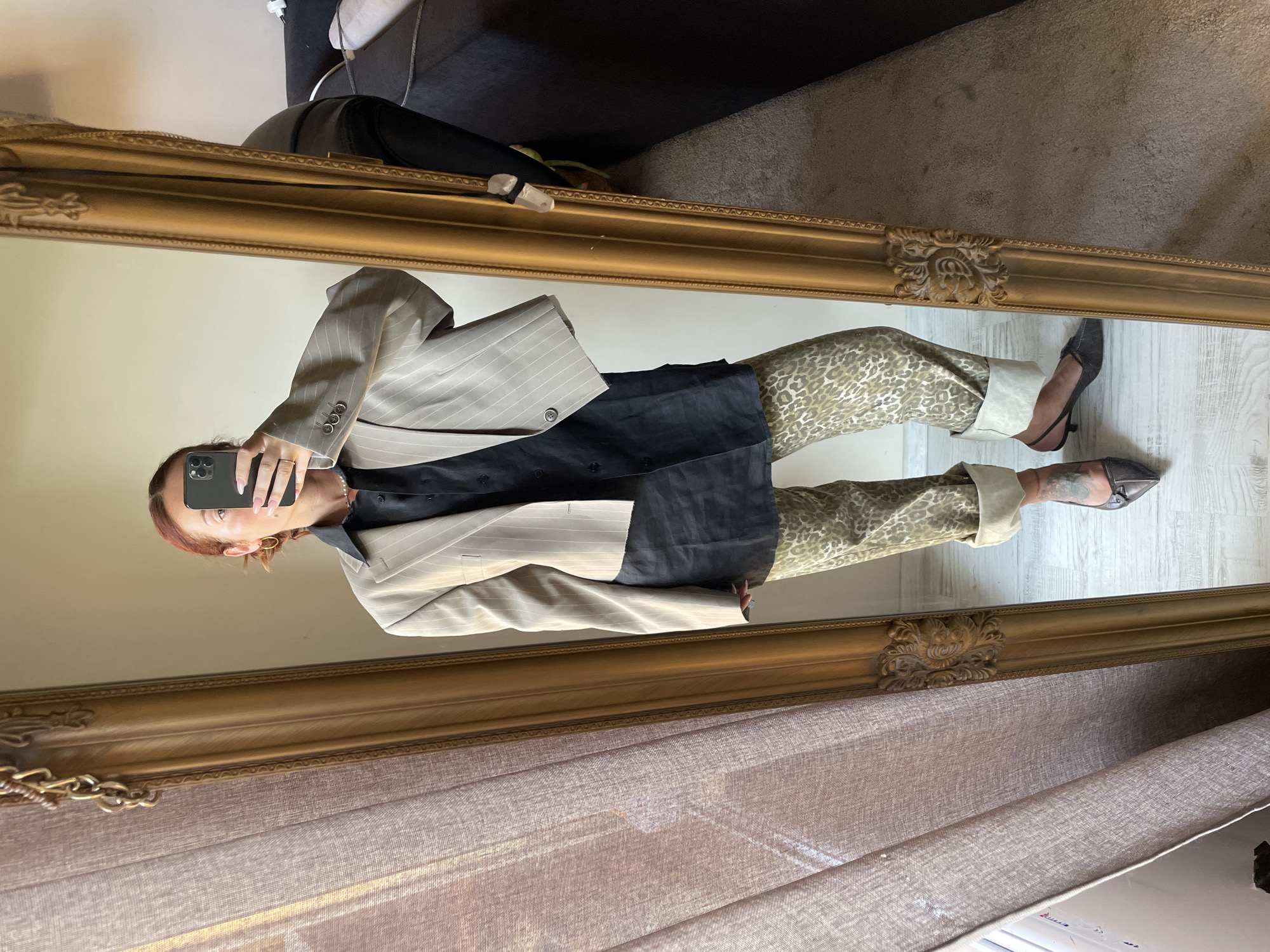 A person is taking a selfie in a mirror, showcasing their outfit. They are wearing a beige pinstripe blazer over a black collared shirt, leopard print pants with cuffed hems, and dark pointed-toe heels. The mirror has an ornate gold frame. The background appears to be an indoor setting, possibly a room in a home, with a curtain to the left and a sofa and carpet visible in the reflection to the right. The flooring is light-colored wood. No specific city or location information is present in the image.