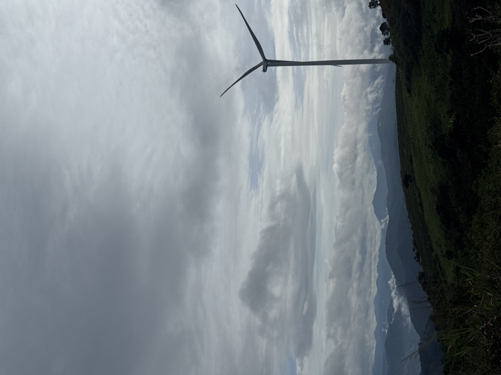 A wind turbine is seen against a cloudy sky in Trường Thọ, Vietnam. The turbine is situated on a green hill that slopes down towards a mountainous landscape shrouded in mist.