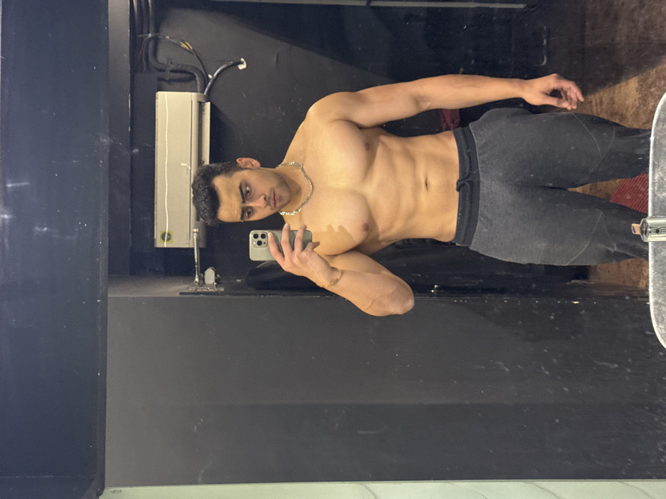 A shirtless man stands in front of a mirror, holding a phone to take a selfie. He is wearing gray sweatpants with a drawstring and a silver chain around his neck. The background is a dimly lit room with dark walls and a visible air conditioning unit. His body is muscular, and he is looking directly at the camera with a slight frown. The overall impression is that of a self-aware fitness enthusiast capturing a moment.