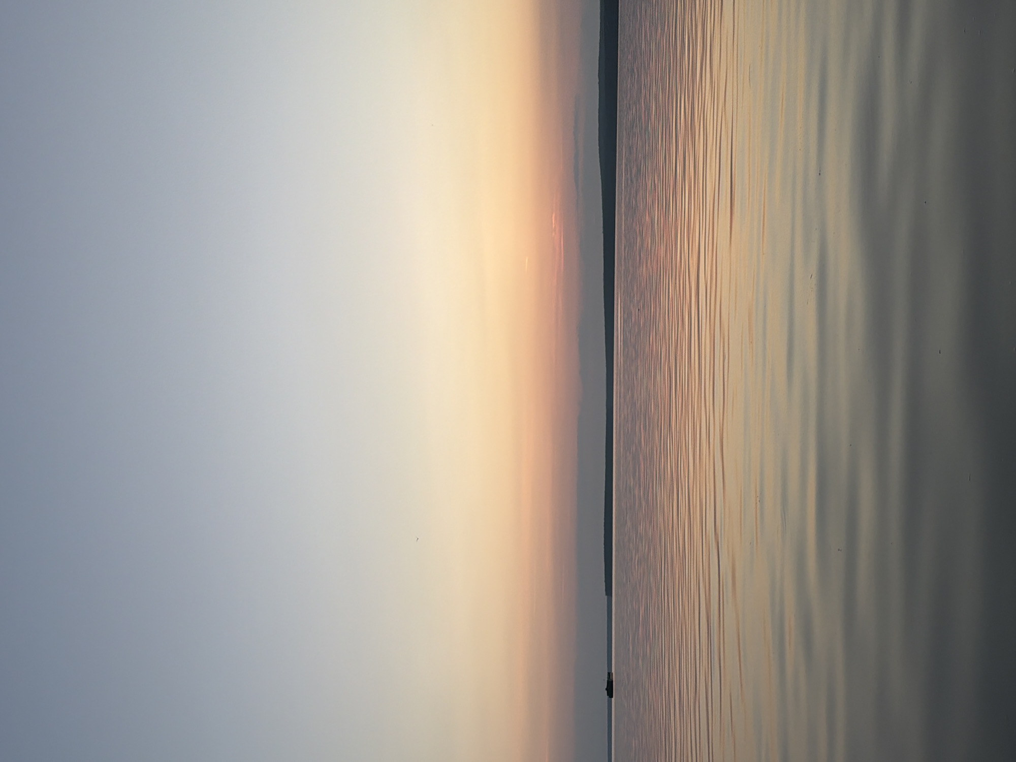 This image depicts a serene sunset over a body of water, likely the Kama River in Chaikovsky, Russia. The sky transitions from a pale blue at the top to soft hues of pink and orange closer to the horizon, indicating the sun has recently set or is about to set. A thin, dark line of trees or land forms the distant horizon. The water's surface is calm, with gentle ripples reflecting the colors of the sky. A small, dark silhouette of a boat is visible on the left side of the frame, positioned on the water near the horizon. The overall atmosphere is peaceful and tranquil.