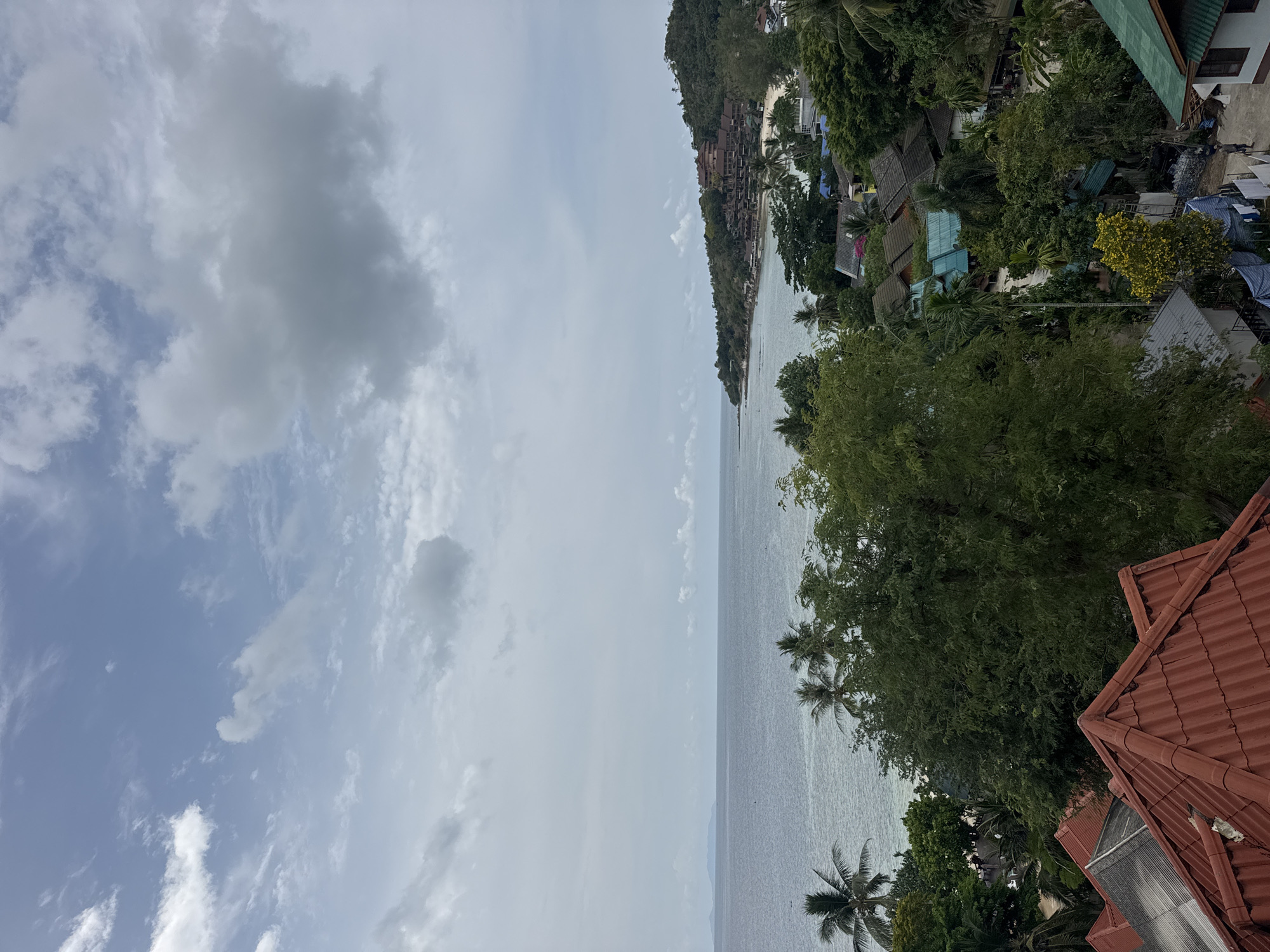 The image depicts a coastal view in Baan Sri Thanu, Thailand, likely taken from an elevated position such as a building rooftop, identified by the visible red roof tiles in the foreground. The scene captures a stretch of coastline with a visible beach and buildings nestled amongst dense green vegetation. The water appears calm. The sky is overcast with varying cloud formations.