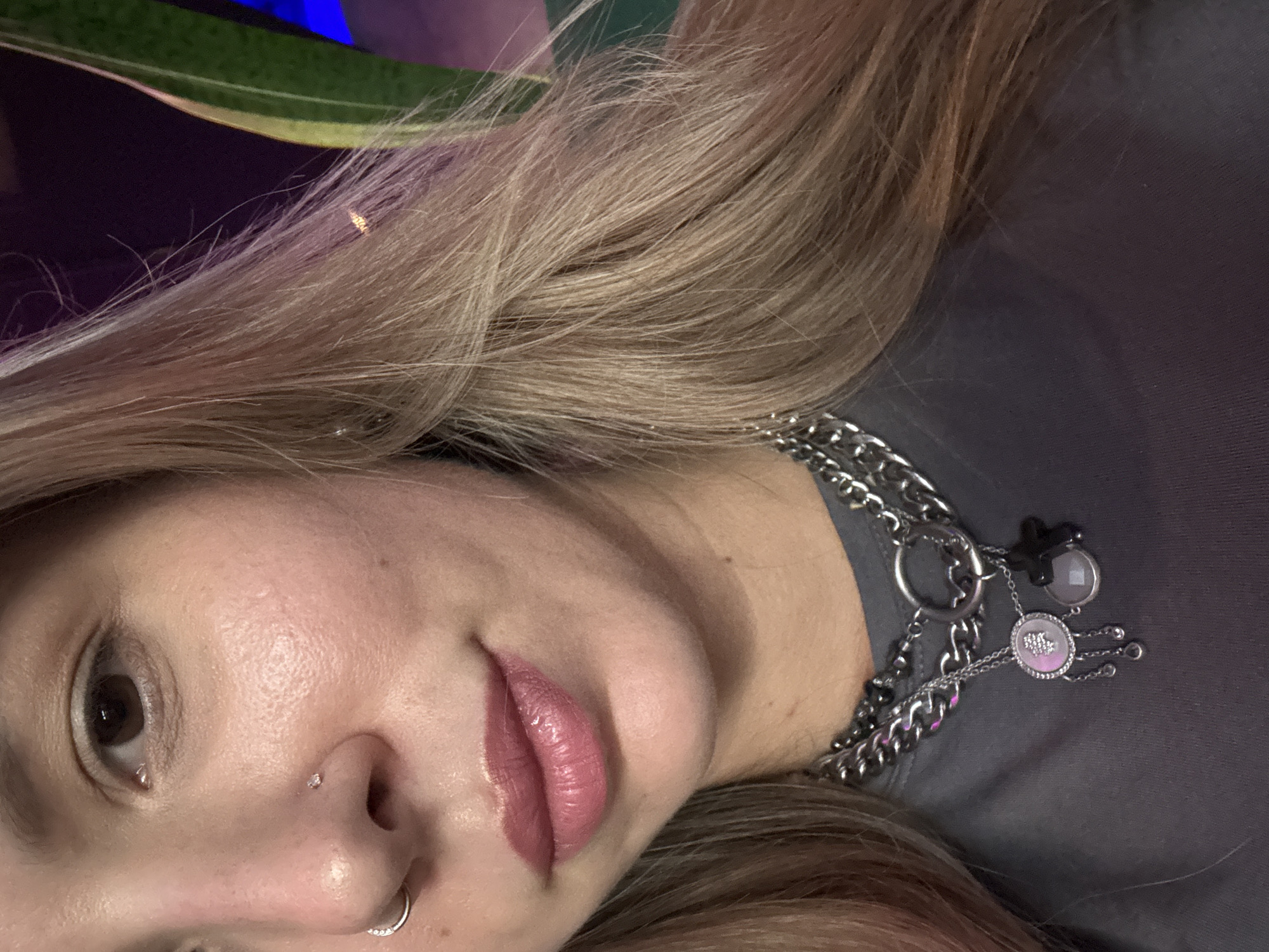 A medium shot shows a close-up of a woman's face and upper chest. She has fair skin, brown eyes, and blonde hair. She is wearing makeup, including lipstick and blush, and has a nose ring and a lip ring. She is also wearing a dark gray t-shirt and two layered silver necklaces, one of which has a pendant with multiple charms. The background is dark and out of focus, with hints of green and purple light. The woman is looking slightly to the left of the camera.