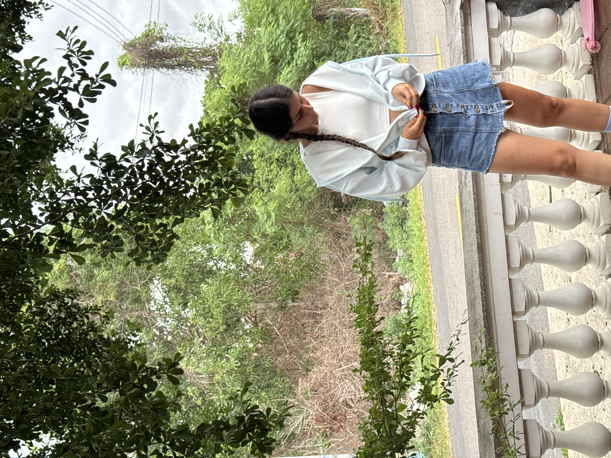A young woman with dark hair braided down her back stands on a white balcony railing. She is wearing a white crop top and a light blue zip-up hoodie, unzipped. Her lower half is clad in a denim skirt with a button-up front. The woman is looking down and appears to be zipping up her hoodie. In the background, lush green trees and vegetation are visible, along with a road and a cloudy sky. A pink scooter is partially visible on the ground to the right. The scene appears to be in a tropical or subtropical climate, likely Mũi Né, Vietnam, given the abundance of greenery and tropical flora.