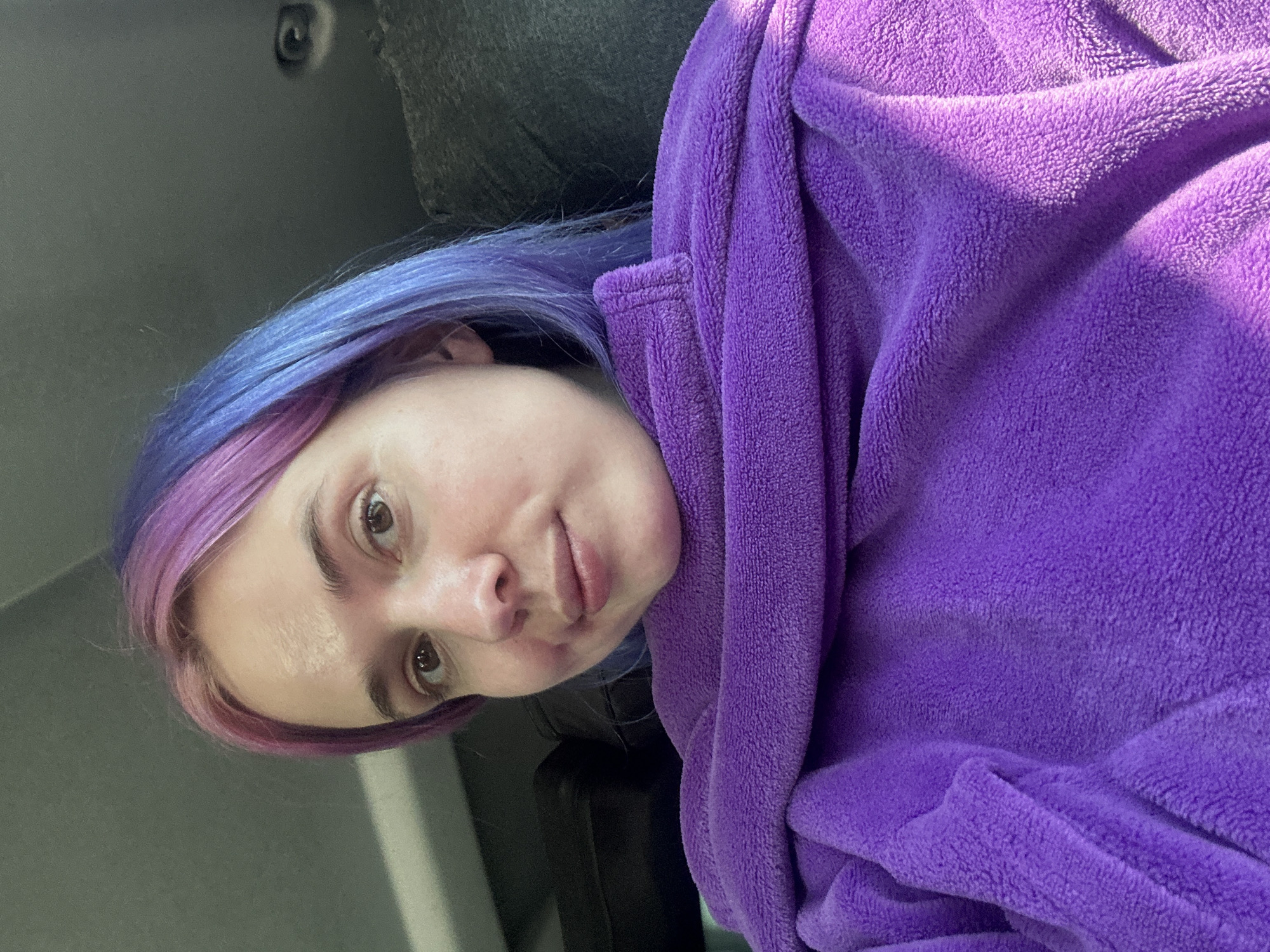 A woman is shown in a close-up, angled portrait. She has light skin, brown eyes, and her hair is dyed in two colors, with the roots near the top of her head colored blue, transitioning to a pink hue towards the tips. She is wrapped in a plush, purple robe. The background is indistinct but suggests an indoor setting. There is no information to suggest this photograph was taken in Le Muy, France.