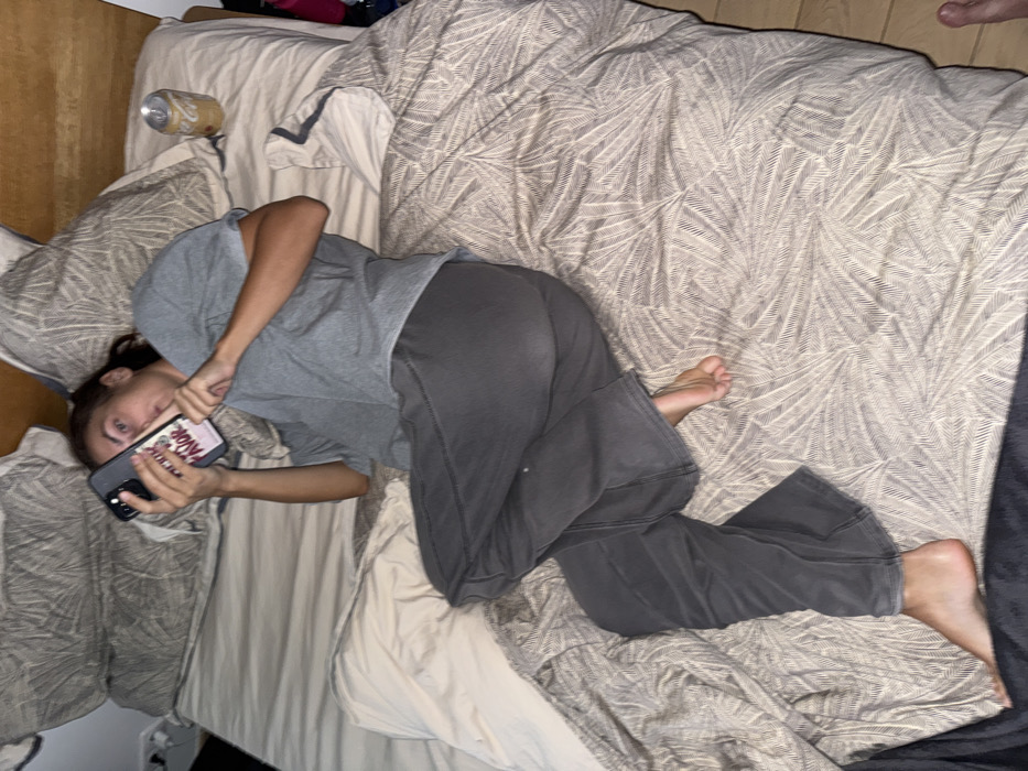 The scene depicts an individual lying on a bed in an indoor setting, consistent with a bedroom. The specific location within the city of Warsaw, Poland, cannot be determined from the visual evidence.
One person, identified as an adult female with dark hair tied back, is positioned on a bed with a light-colored wooden headboard. She is lying on her back, angled towards the left, with her legs bent and crossed, exposing bare feet. She is dressed in a short-sleeved grey t-shirt and dark grey pants. Both hands hold a dark-colored smartphone horizontally in front of her face; the phone case is light pink with dark text, partially legible as "FAVOR". Her eyes are wide, and her mouth is slightly open, with her gaze directed towards the camera. To the upper right of her head, on the patterned duvet cover, rests an open, light gold or silver-colored aluminum beverage can. The bed is covered with a light-toned duvet featuring a repetitive, leaf-like pattern, and includes two grey patterned pillows and one white pillow. The environment is dimly lit.
