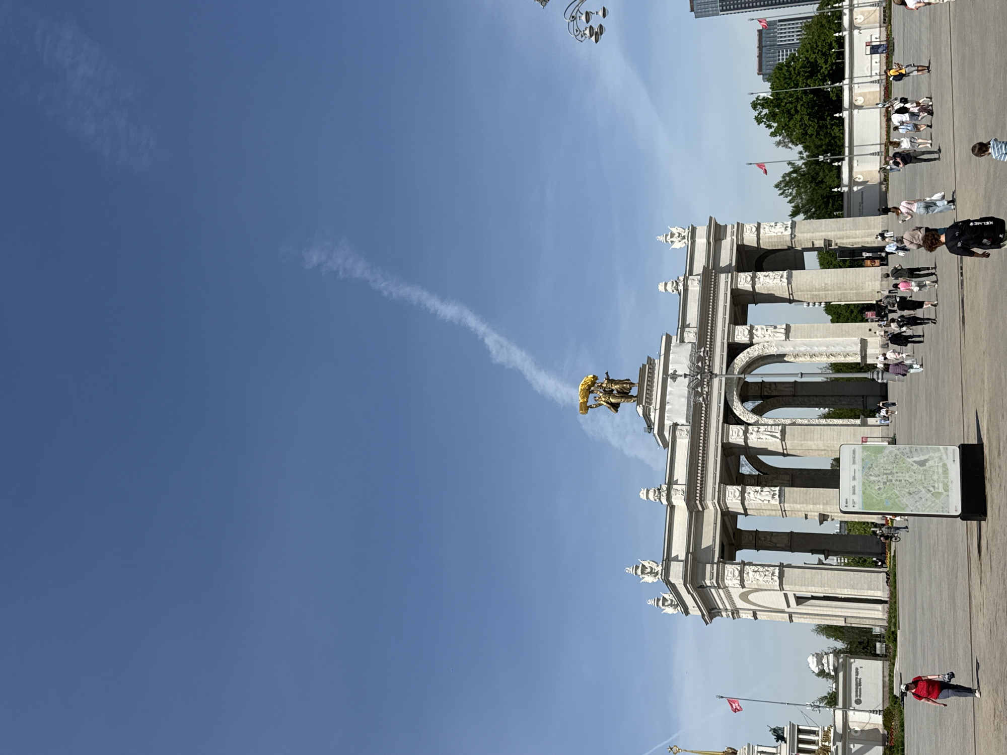 The image depicts an urban landscape with a clear blue sky dominating a significant portion of the frame. A contrail stretches across the sky. The primary architectural feature is a grand triumphal arch, constructed of white stone and decorated with intricate sculptures. Atop the arch is a golden statue. People are milling around the base of the arch, suggesting it is a popular landmark. Also near the arch is a map. The urban setting is confirmed by the presence of buildings in the background. The location appears to be Victory Park in Moscow, Russia.