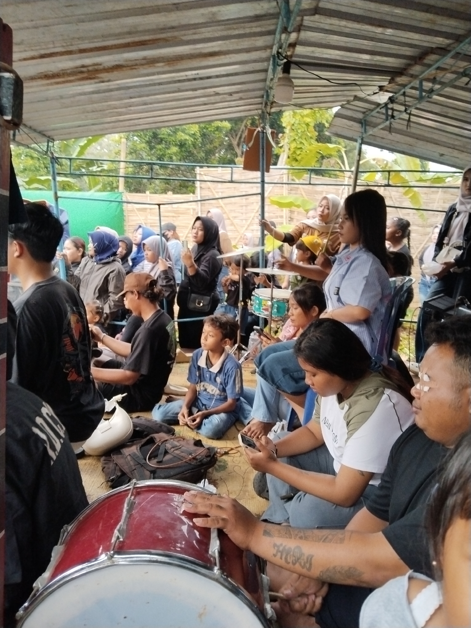A crowd of people are gathered under a corrugated metal roof structure, presumably in Kuto, Indonesia. The crowd includes men, women, and children of varying ages, and some women wear hijabs. Some individuals are seated on the floor while others are in chairs. A drum set is prominent in the foreground, with a person’s hand resting on a large red drum. One woman sits in a chair and appears to be looking at the drum set as well. A woman is holding up a drumstick as if she is about to play the drums, and a small child sits in front of her on what appears to be the lap of another woman. Many appear to be watching the drum-related activity. Trees and a bamboo fence are visible in the background.