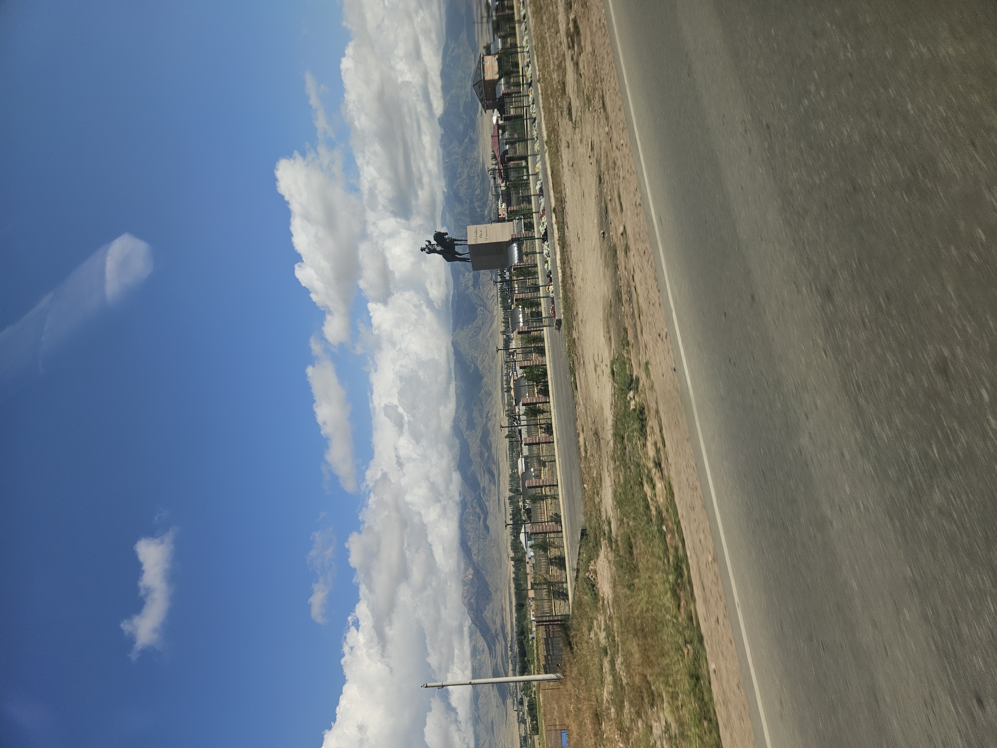 The image captures a vast landscape under a bright blue sky filled with scattered white clouds. In the middle ground, a prominent equestrian statue stands on a raised platform, depicting a person on horseback. The statue is positioned in front of a fence, behind which several buildings, possibly houses, are visible. Further in the background, a range of mountains stretches across the horizon, with some appearing snow-capped. The foreground is dominated by a paved road, suggesting a view from a moving vehicle. The environment appears to be arid, with dry vegetation and sparse trees visible along the roadside and around the buildings. The overall scene suggests an outdoor setting in a semi-urban or rural area, possibly with a historical or commemorative significance due to the presence of the statue. The exact location in the city of Ornek, Kyrgyzstan, is not explicitly identified in the image, but the context suggests a location within or on the outskirts of the city.