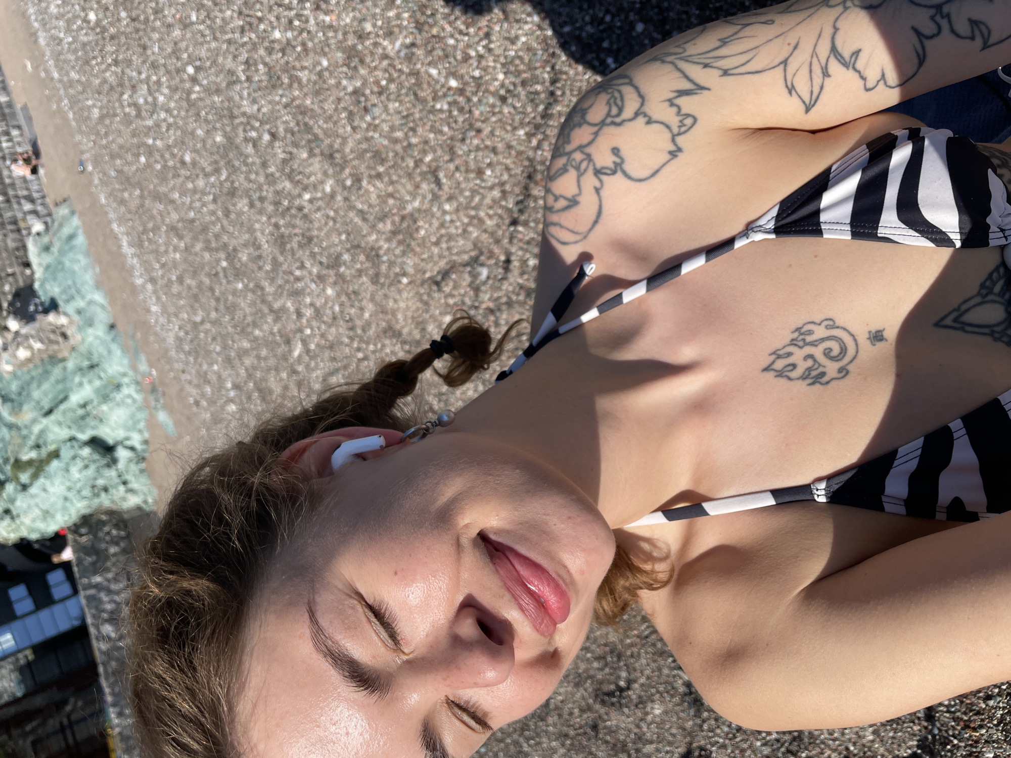 A young woman lies on a beach in Rafailovići, Montenegro. She wears a black and white striped bikini top, and she has several tattoos on her upper body. She has light brown hair pulled back in a braid and wears a white earbud in one ear. Her eyes are closed, and she appears to be sunbathing. The beach consists of small pebbles and stones. In the background, there is a building. The image is taken from a high angle, looking down on the woman.