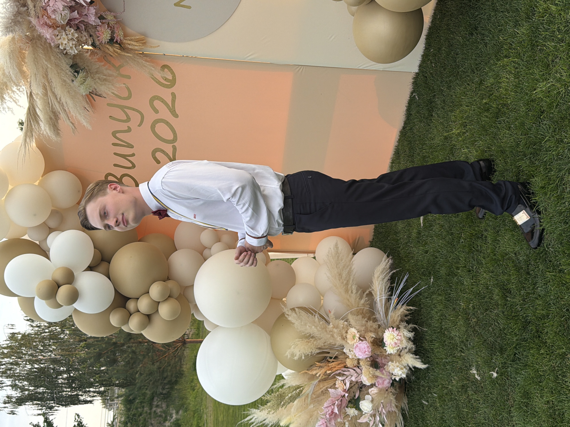 A young man stands outdoors in front of a backdrop decorated with balloons and dried floral arrangements. He is wearing a white dress shirt, dark pants, and a bow tie. The backdrop features the text "Bunyak 2026" in gold lettering. The scene appears to be set up for a celebration or event, possibly a graduation or formal gathering. The time of day is likely afternoon, as suggested by the lighting. The location is Khotianivka, Ukraine. The man's expression is neutral.