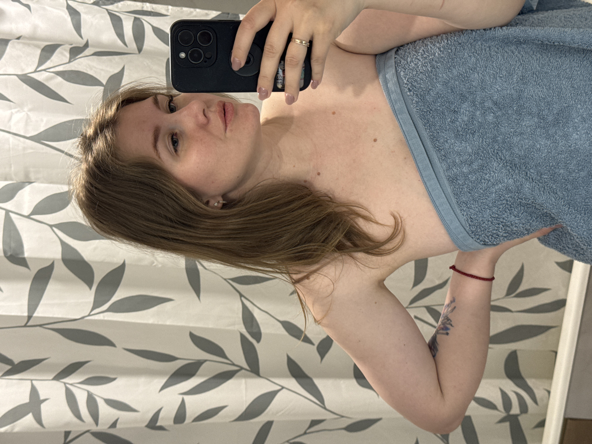 A woman is taking a selfie in a bathroom. She is wrapped in a blue towel. She is holding a black smartphone in her right hand. The background shows a white shower curtain with grey leaf patterns. The location is Gornyi (Dvurechenskoe S/P), Russia.