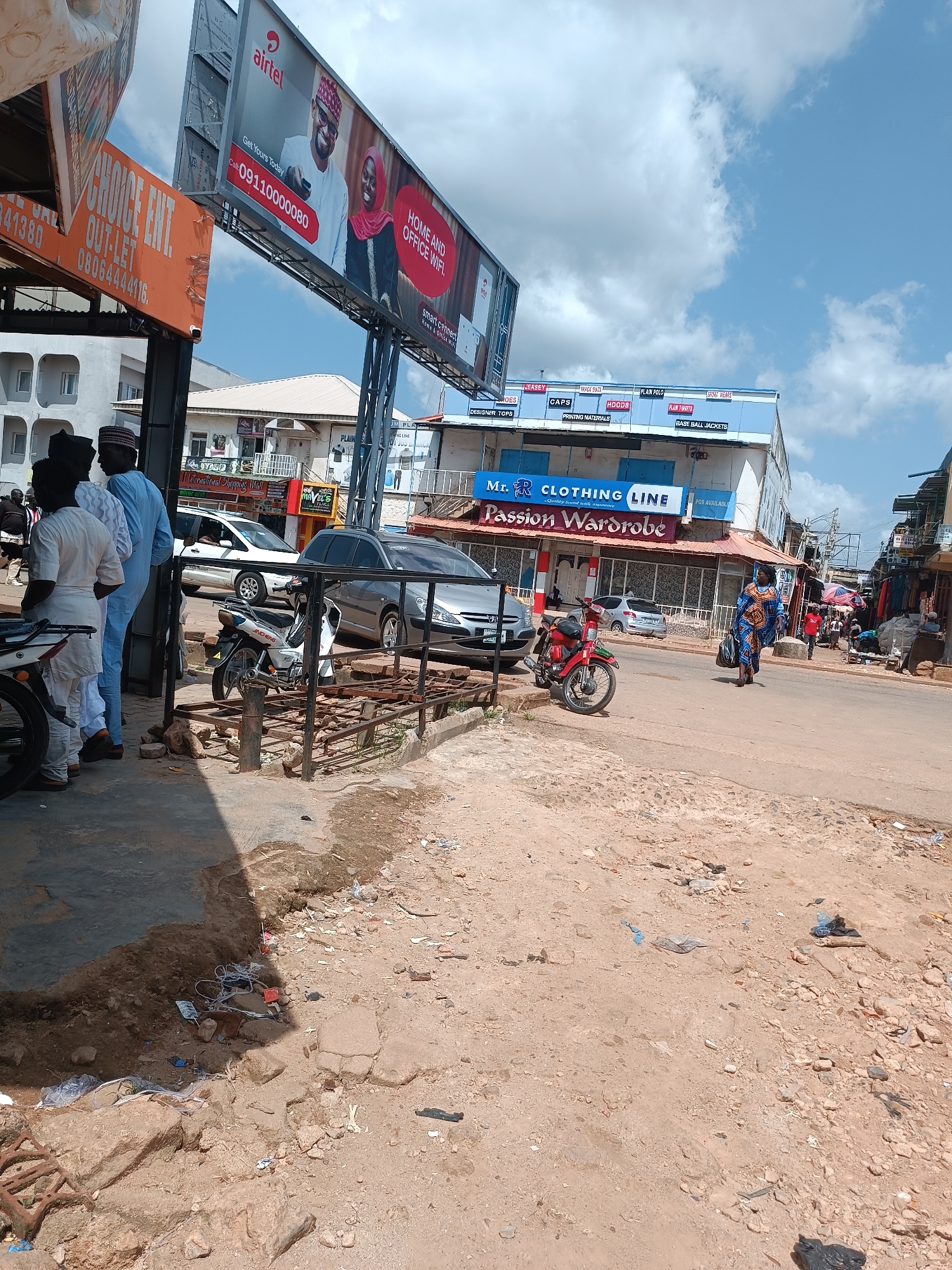 This is a street-level photograph taken in Jos, Nigeria, on a clear day with a few clouds in the sky. The scene depicts a busy commercial area with several buildings and various elements characteristic of urban Nigerian street life.
In the foreground, on the left, a building facade is partially visible, with an orange sign that reads "CHOICE ENT. OUT-LET" and a phone number. A shadow falls across the ground, revealing scattered debris, including plastic bags, wires, and broken concrete.
Dominating the upper portion of the image is a large billboard advertising "airtel" with images of a man and a woman, along with the text "HOME AND OFFICE WIFI." Below this billboard, another set of signs for businesses is visible. One sign reads "Mr. R CLOTHING LINE" and below it "Passion Wardrobe." Other smaller signs indicate clothing items and services.
The street itself is paved, with a few vehicles present. A white motorcycle is parked near the foreground, and a red motorcycle is further down the street. A silver car and a grey car are also visible, suggesting traffic.
Several people are present in the scene. On the left, three men stand near the building facade, two of whom are wearing traditional attire and hats. Further down the street, a woman in a colorful patterned dress walks carrying bags. Other figures are seen in the background, some appearing to be shopkeepers or customers.
The environment appears somewhat unkempt, with debris and uneven ground visible in several areas, particularly in the foreground and along the side of the street. The buildings are of varying designs, with awnings and shopfronts visible. Overhead, electrical wires and poles suggest a functioning urban infrastructure. The overall impression is one of a vibrant, bustling marketplace environment.