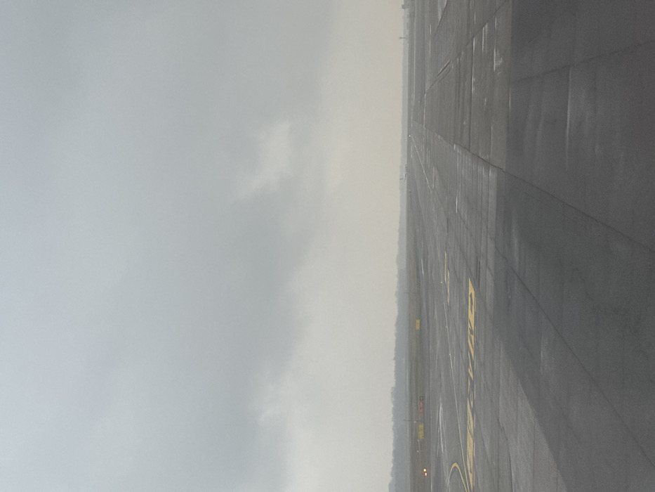 The scene depicts a section of an airport tarmac at Milan Malpensa Airport (MXP) in Somma Lombardo, Italy, under an overcast sky. The dark grey asphalt surface is wet, indicating recent precipitation, and features numerous yellow painted markings, including directional arrows, lines, and alphanumeric identifiers such as "A2" and "CM". In the mid-distance on the left, several orange-red airfield lights are illuminated along the edge of the tarmac. Further down the runway, a white aircraft, likely an airliner, is visible with its front lights illuminated. A hazy, dark green tree line stretches across the horizon, separating the airfield from the background. A faint, vertical structure, possibly a control tower or airport building, is minimally visible on the far right horizon. No individuals are present.