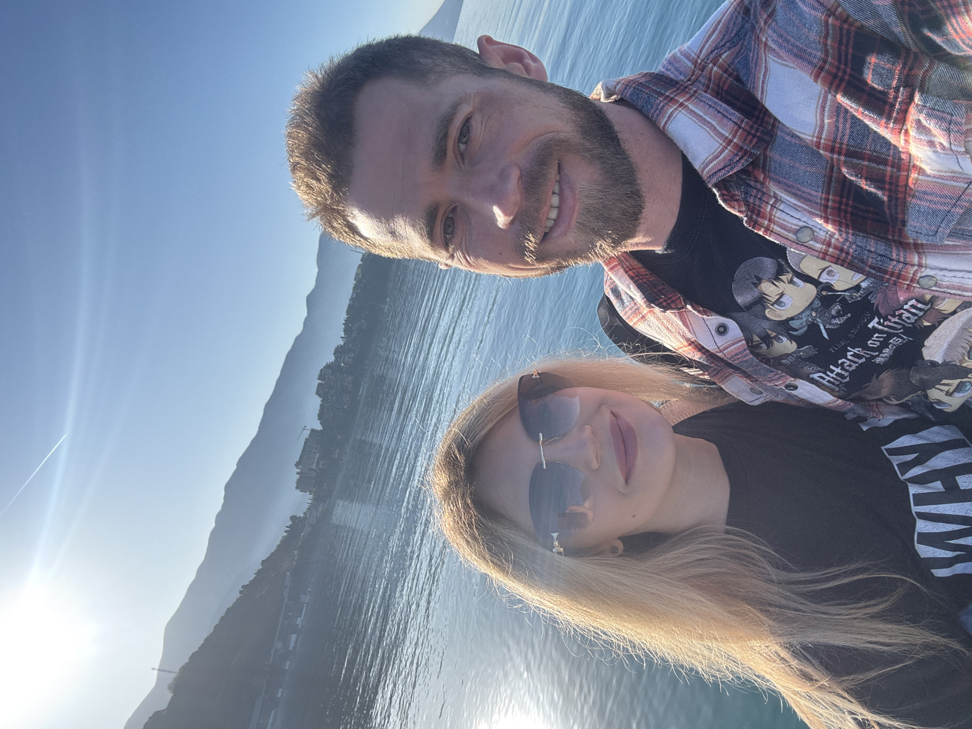 The image shows a selfie of a man and a woman, presumably a couple, taken outdoors. The man is in the upper part of the frame, wearing a red and white flannel shirt over a black t-shirt with "Attack on Titan" graphic. He has short brown hair and a beard. The woman is below him, wearing sunglasses and a black t-shirt. She has long blonde hair. They are both smiling.
The background reveals a waterfront scene. There is a body of water, likely the Adriatic Sea, with mountains visible in the distance. Buildings are visible along the shoreline, suggesting a city or town. The light suggests it's either early morning or late afternoon, with the sun low in the sky. While the exact location within Budva is not explicitly discernable from the image alone, the landscape is consistent with a coastal setting in Montenegro, specifically the Budva Riviera.